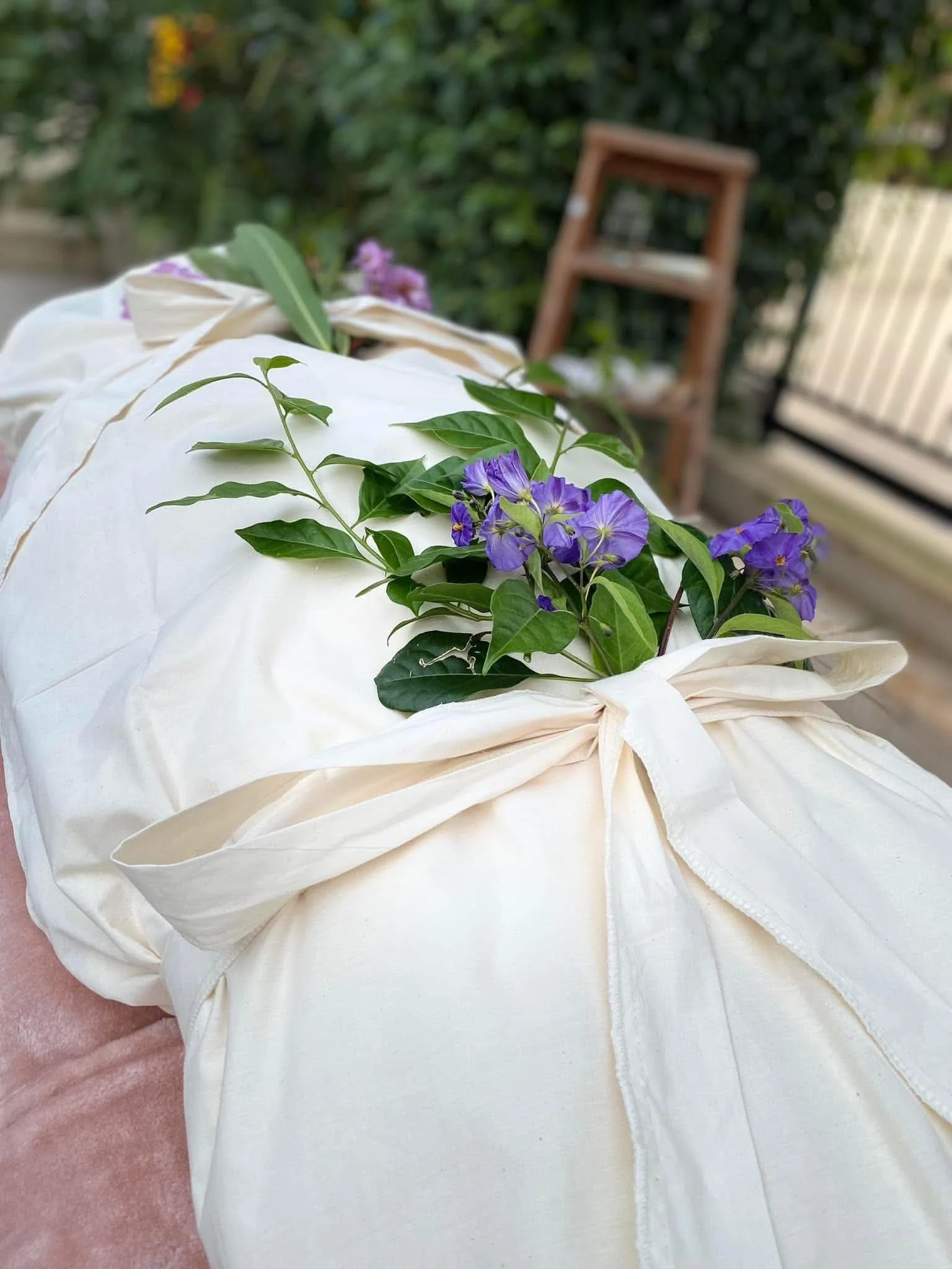 A white fabric bag filled with purple flowers and green leaves, placed on a pinkish surface outdoors with a garden, wooden ladder, and a white fence in the background.
