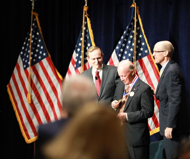 People at an event with American flags in the background, one man wearing a medal.