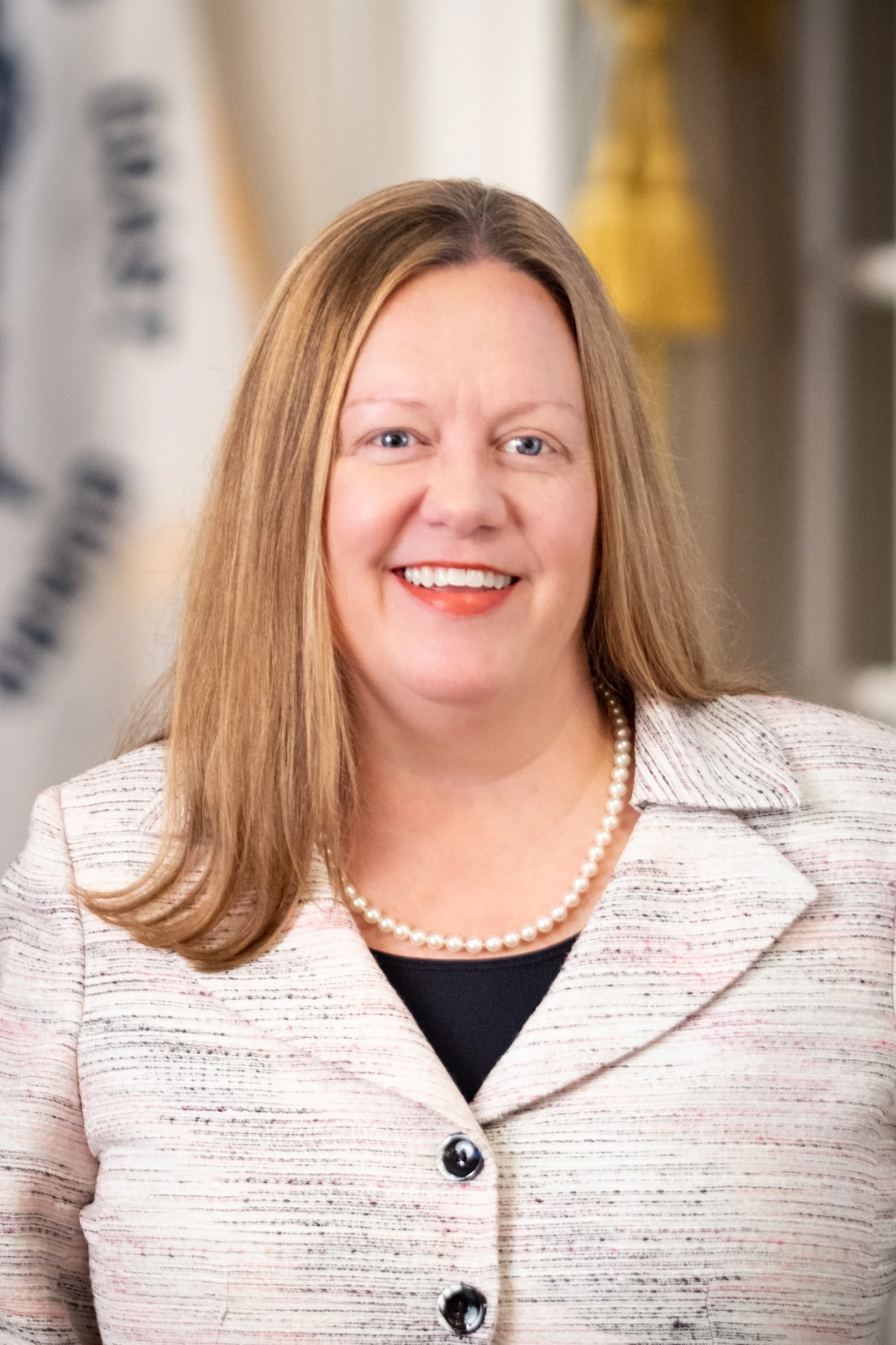 Portrait of a woman with long blonde hair and a black blazer, smiling at the camera against a gray background.