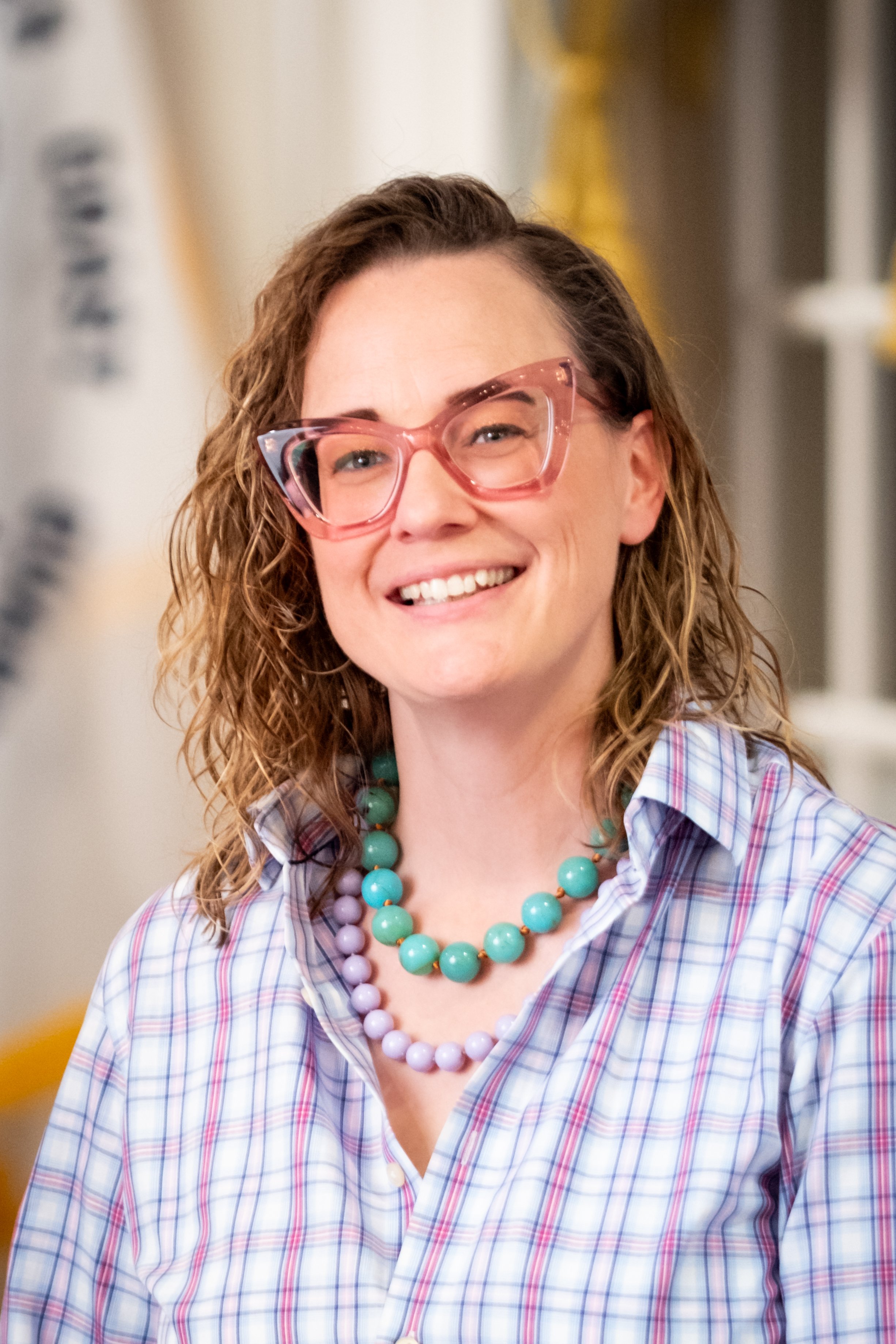 Person with short, curly hair, wearing green glasses and a navy top, smiling in front of a textured concrete wall.