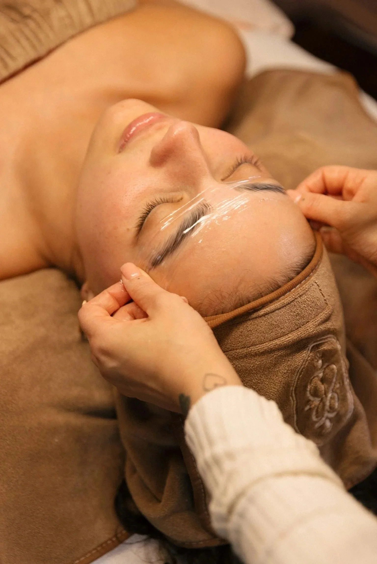 A woman is lying down with her eyes closed, receiving an eyelash extension treatment from a professional. The professional is carefully attaching false eyelashes to her natural lashes.