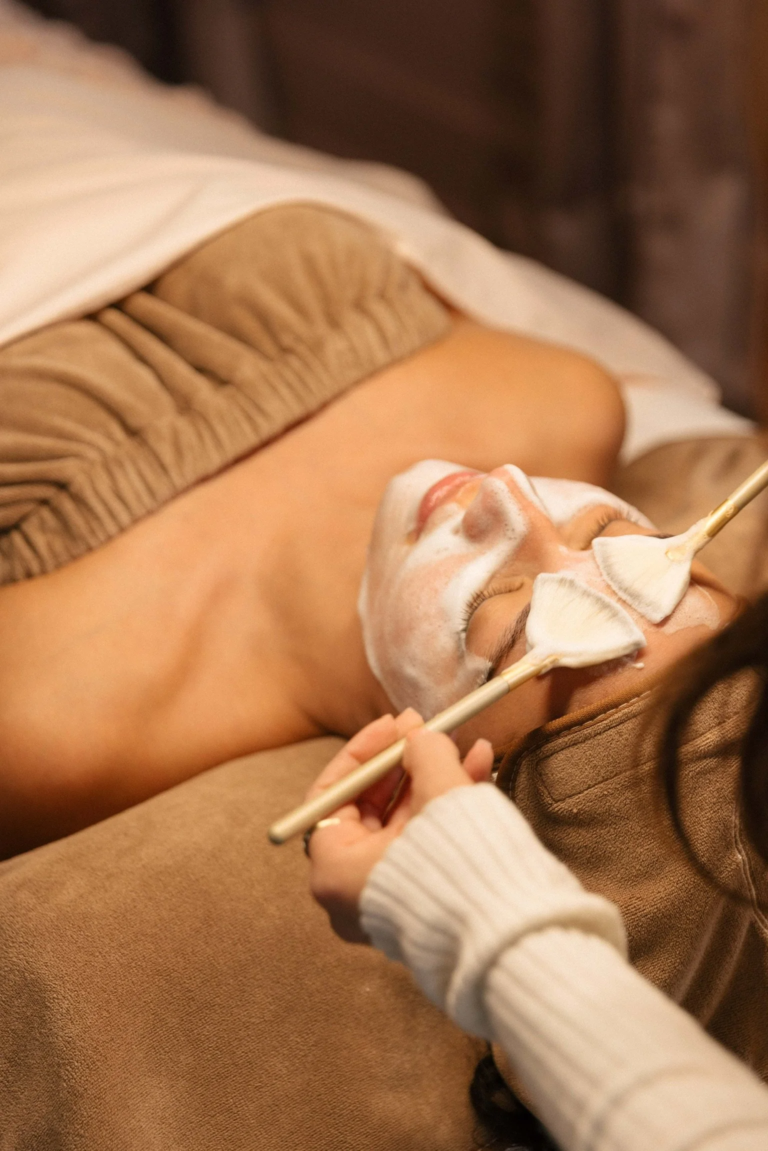 A woman is lying down with her eyes closed, receiving a facial treatment. Her face is covered in a foamy mask, and a skincare therapist is using a brush and a fan-shaped applicator to apply a product on her face. The woman is relaxed, and the setting appears to be a spa or美容spa.
