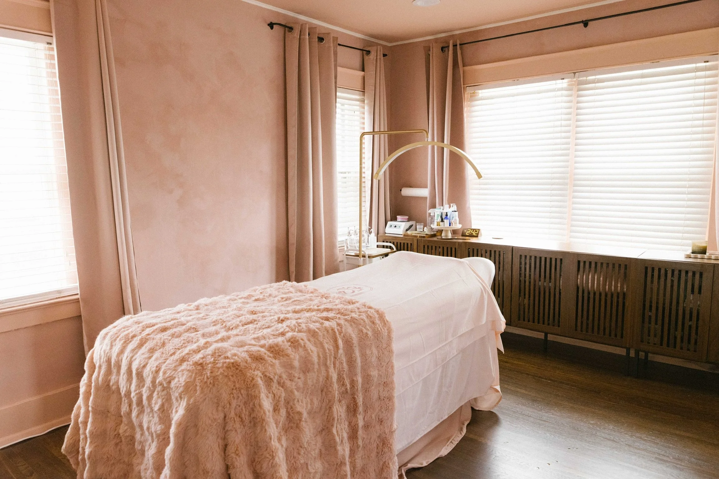 Hotel room with a massage bed covered in a pink blanket, beige curtains on windows, wooden floor, and a cabinet with medical supplies and a digital clock showing 2:42.