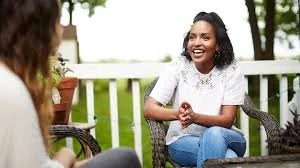 Two women sitting outside on a porch, engaging in conversation and smiling.