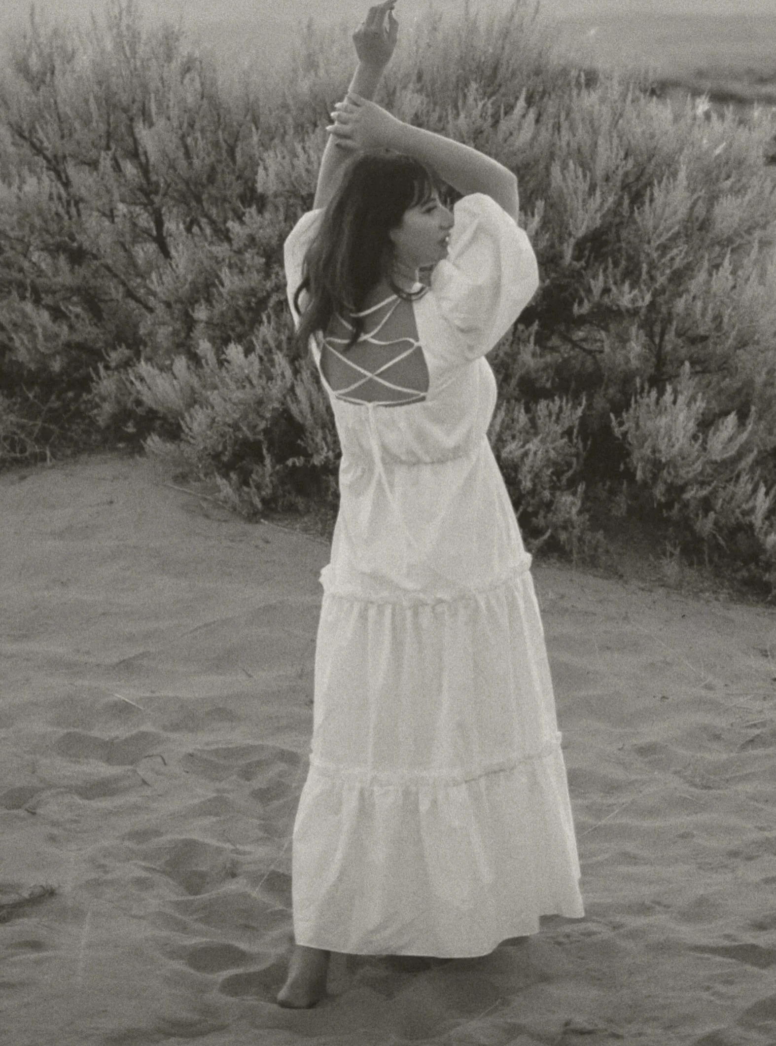 A woman in a long, white dress with puffed sleeves and a crisscross back design, standing barefoot on sandy ground outdoors. She is stretching her arms above her head, with one hand clasping the other. The background features bushy plants and a distant landscape, suggesting a natural setting.