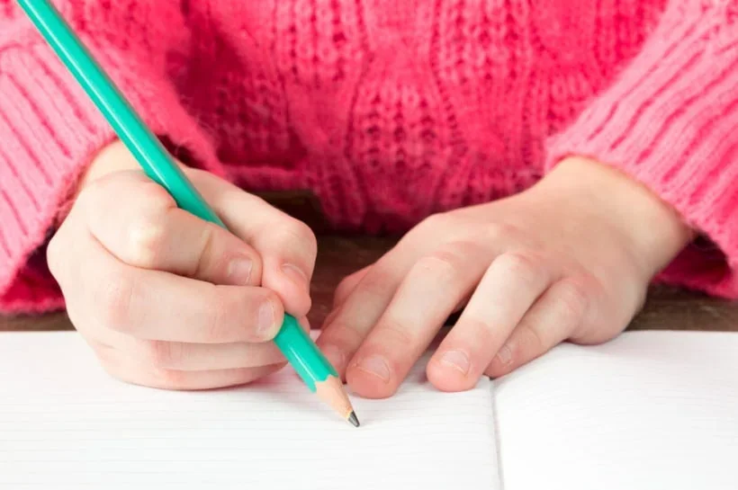 A person wearing a pink sweater writing with a teal pen in a notebook.