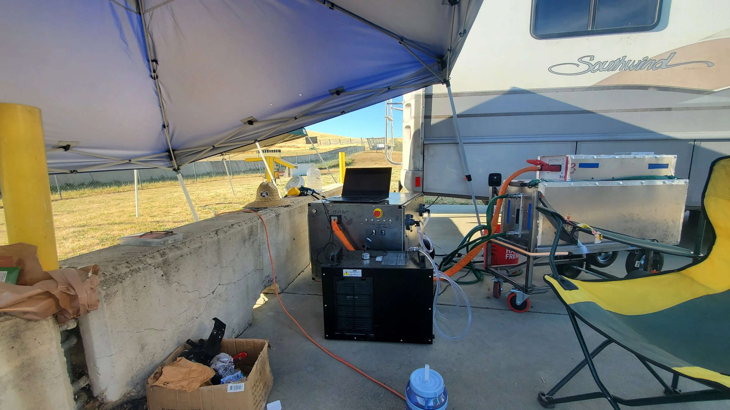 Setup of electric vehicle charging station outdoors under a canopy, with batteries, cables, and equipment connected on a concrete surface.