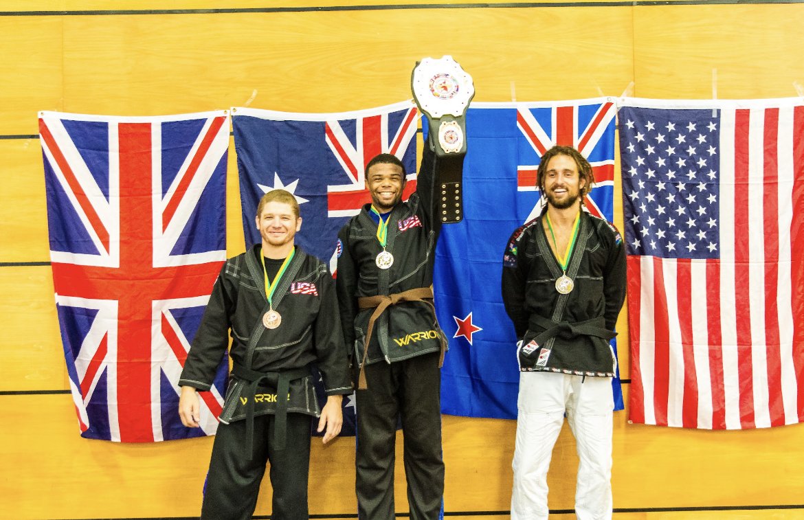 Three martial artists on a podium with medals, in front of British, Australian, New Zealand, and American flags. The central person holds a championship belt.