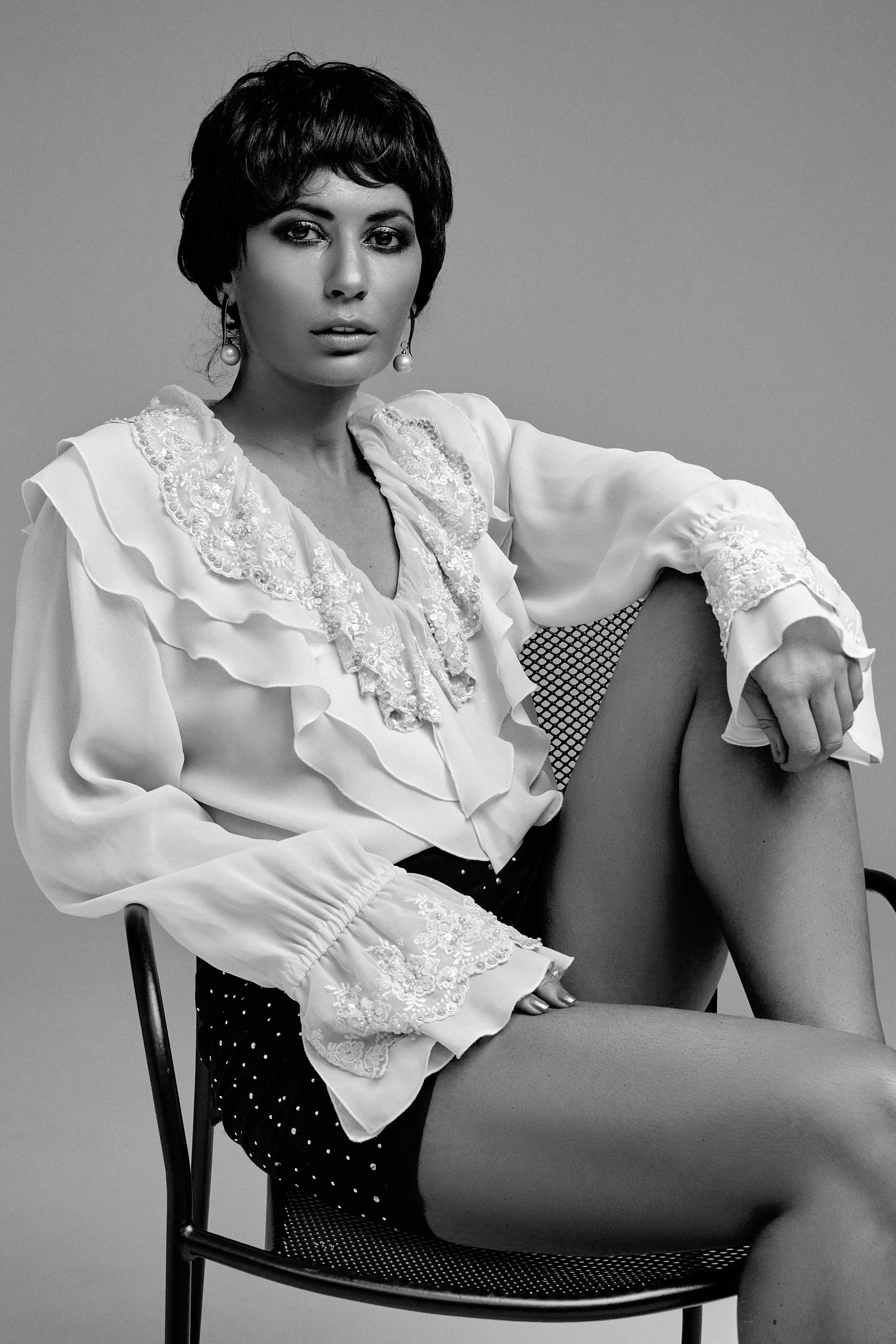 Black and white portrait of a woman wearing a white ruffled blouse, sitting on a chair with a thoughtful expression.