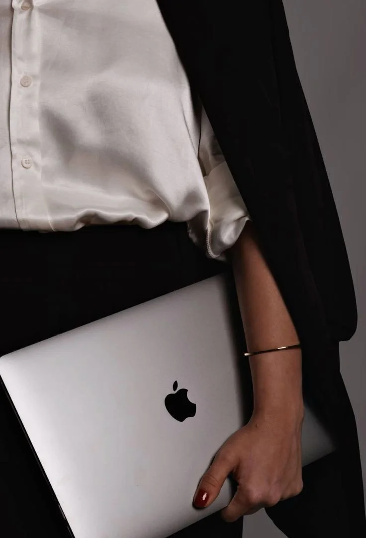 Person holding a MacBook with Apple logo, dressed in a white blouse and black suit.