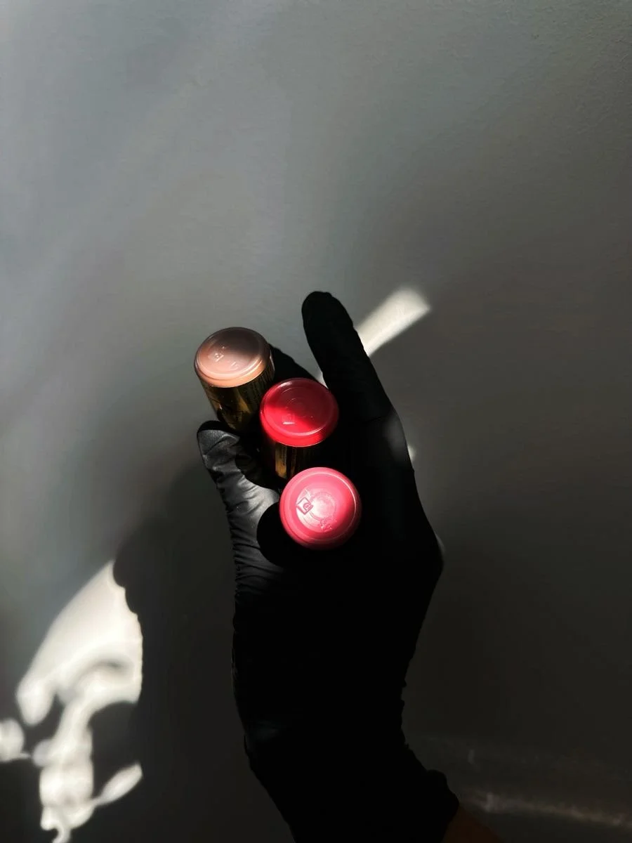 Person holding three lipstick tubes wearing a black glove against a gray background.