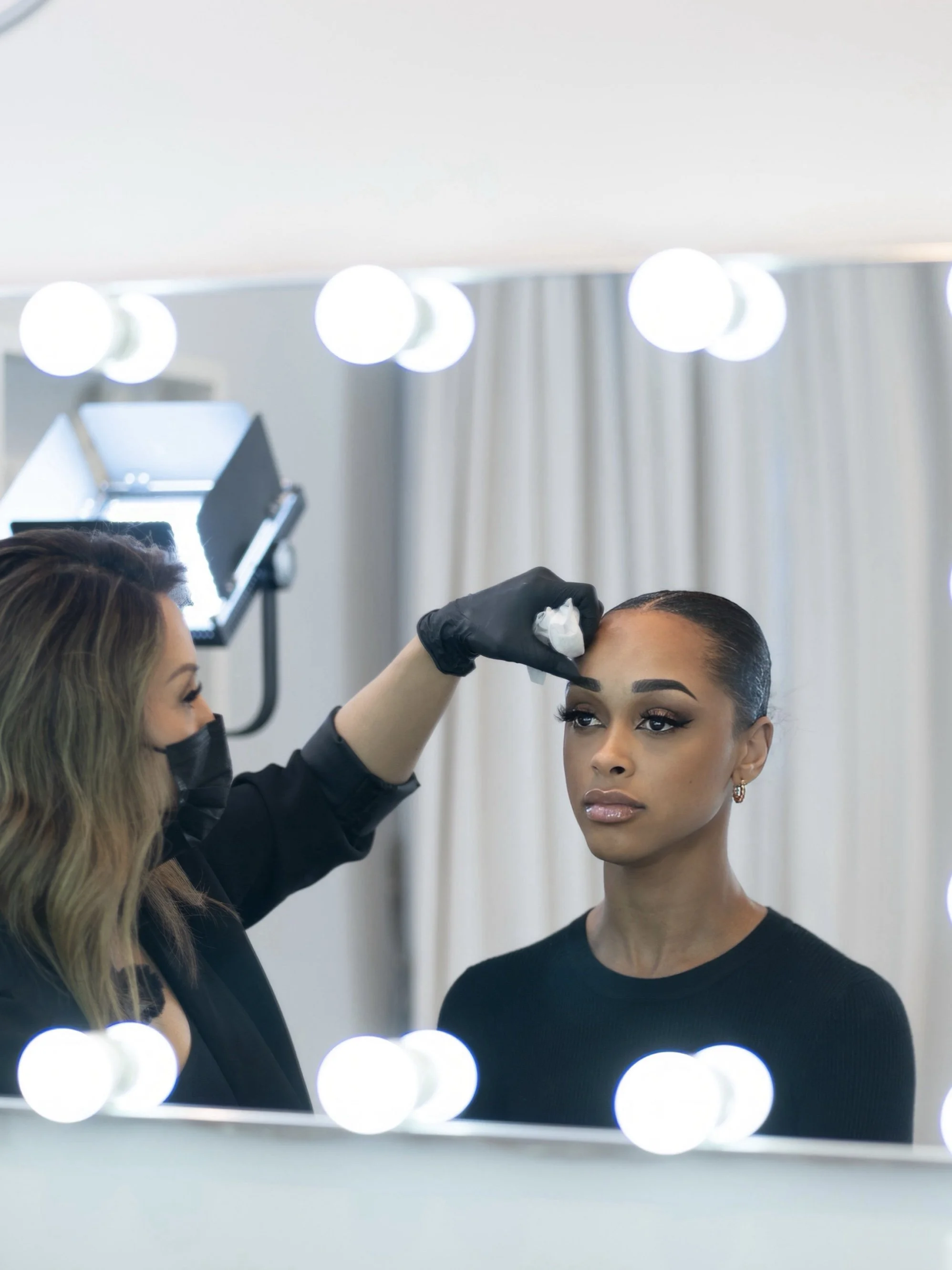 Makeup artist applying makeup to woman in front of a mirror with lights