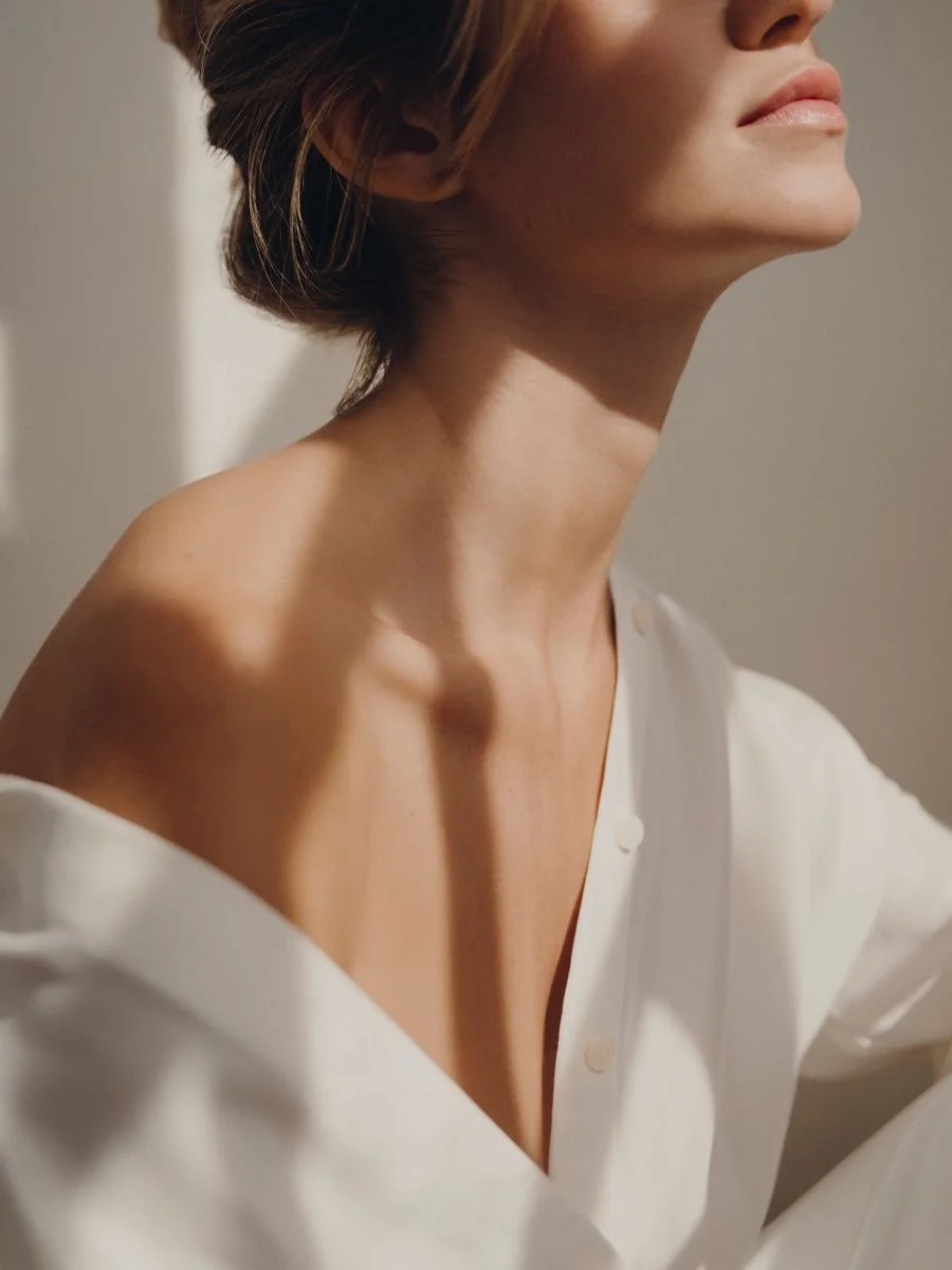 Close-up of a woman's neck and shoulder, wearing a white, off-shoulder shirt, with natural light and soft shadows.