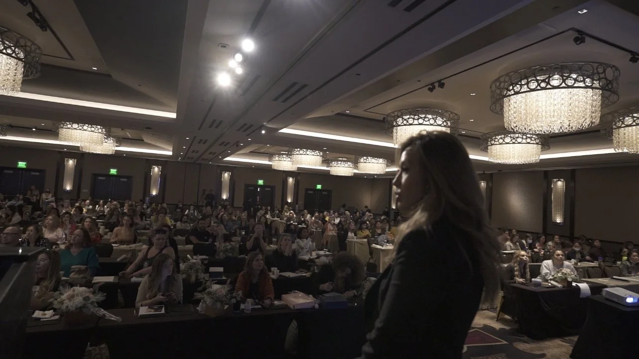 Woman speaking at a conference in a large ballroom with an audience seated at tables, featuring elegant chandeliers overhead.