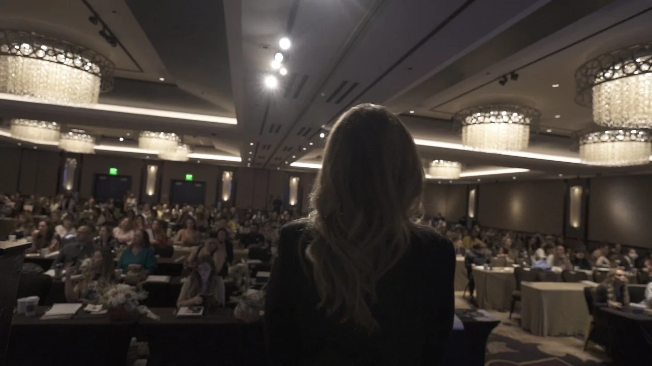 Speaker addressing an audience in a large conference room with chandeliers.