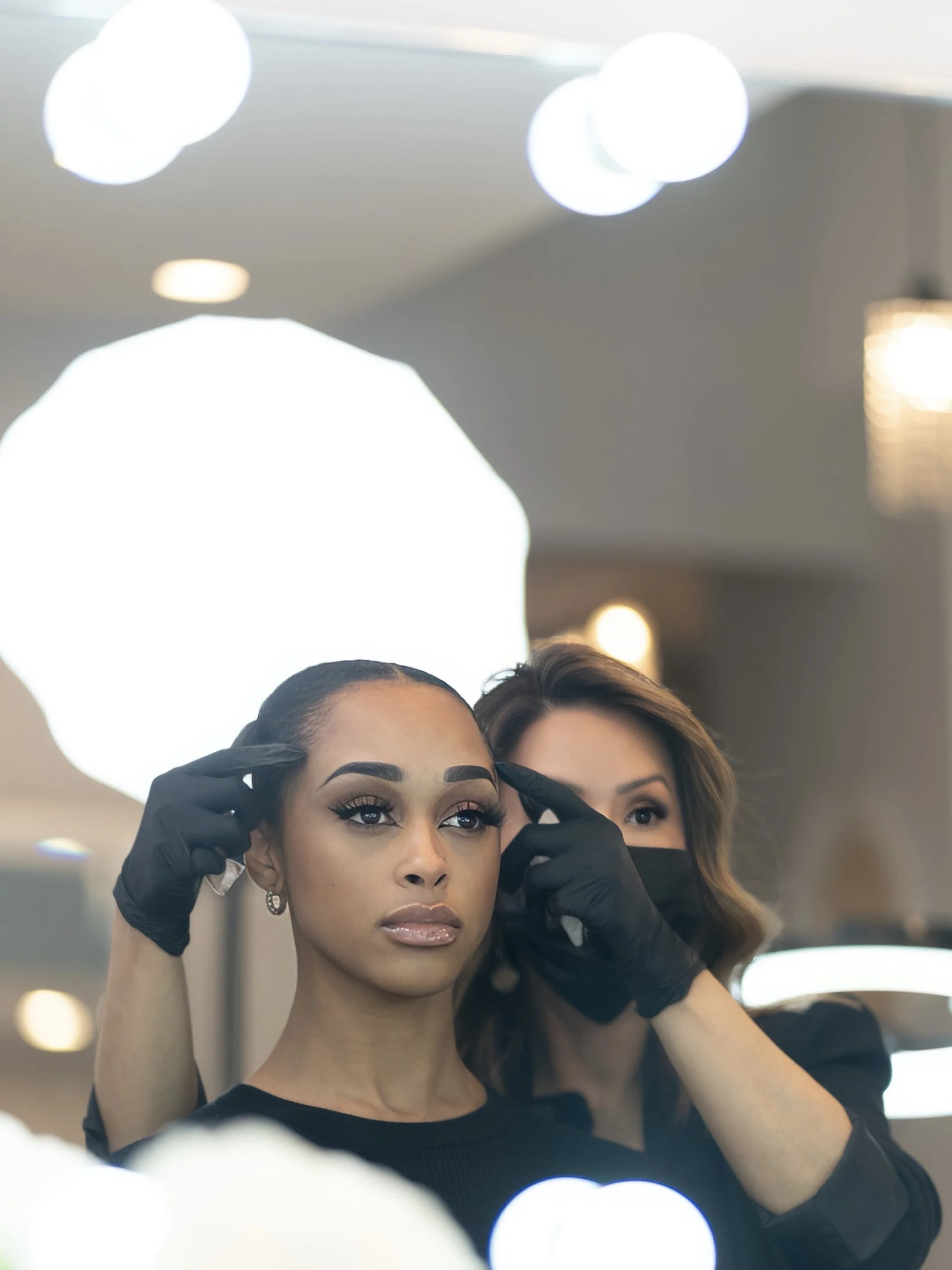 Makeup artist applying makeup to a woman's face in front of a mirror with bright lights.