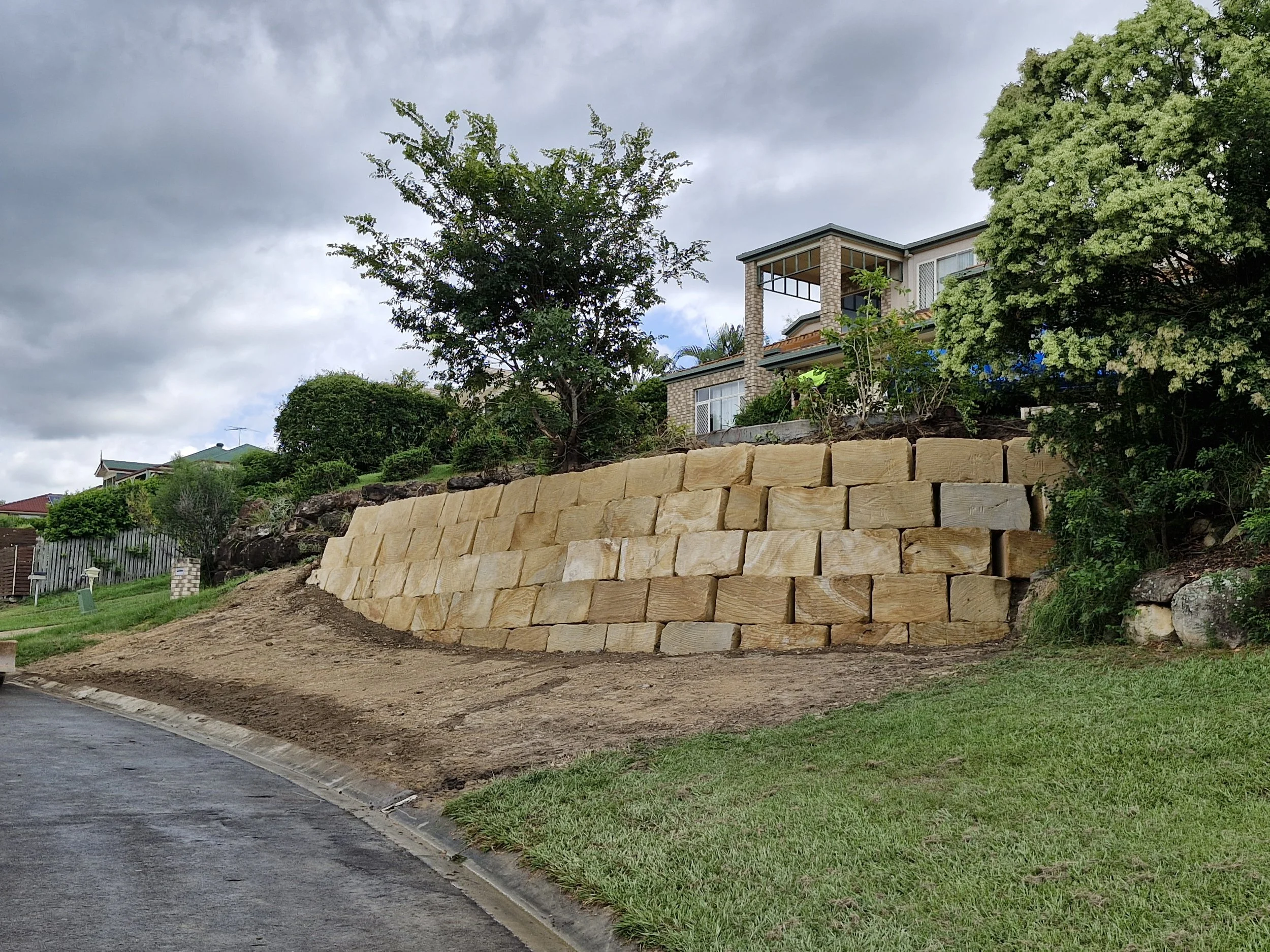 A retaining wall made of large beige stones built on a sloped yard in front of a house, with greenery and trees around, and a cloudy sky overhead.