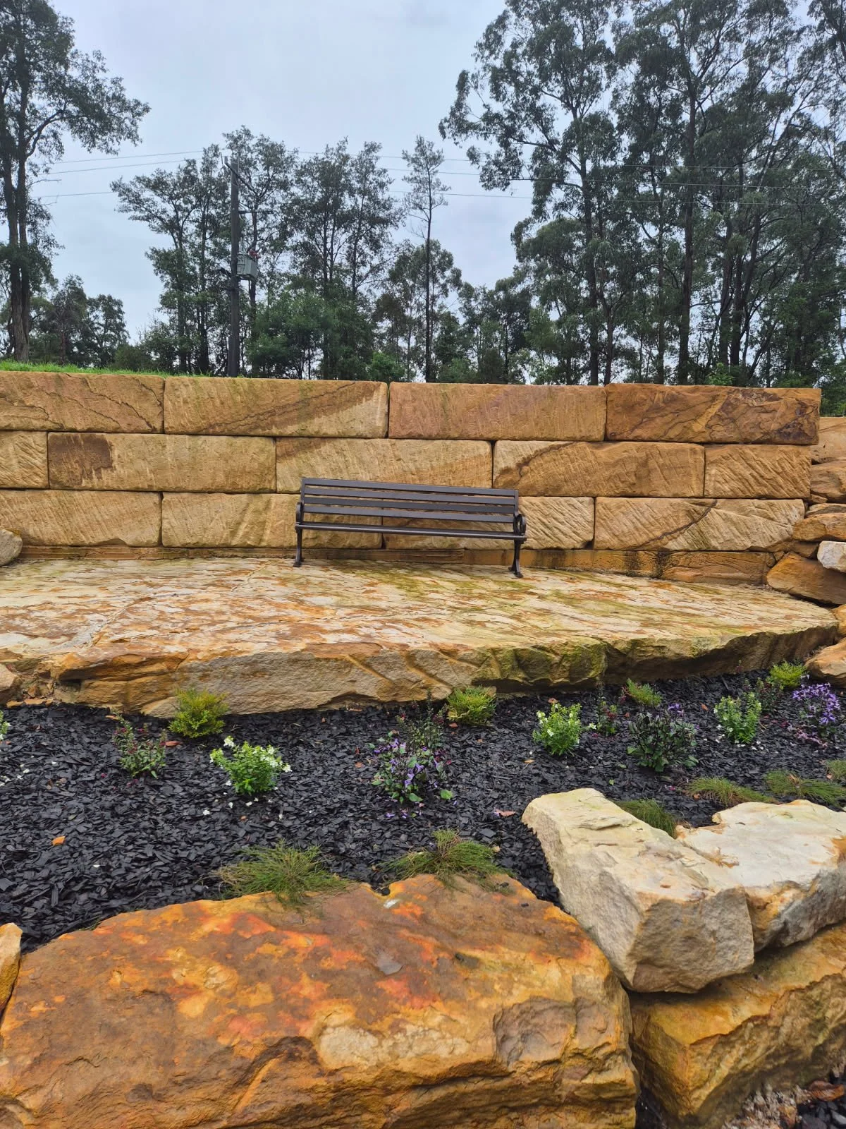 A landscaped outdoor area with a stone wall, a bench, and small plants and flowers in a flowerbed.