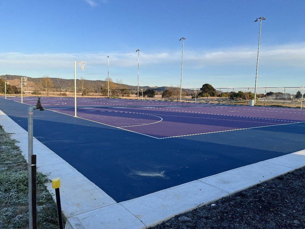 An outdoor basketball court with purple and blue surfaces, surrounded by tall lights and a fence, set against a backdrop of hills and a clear blue sky.