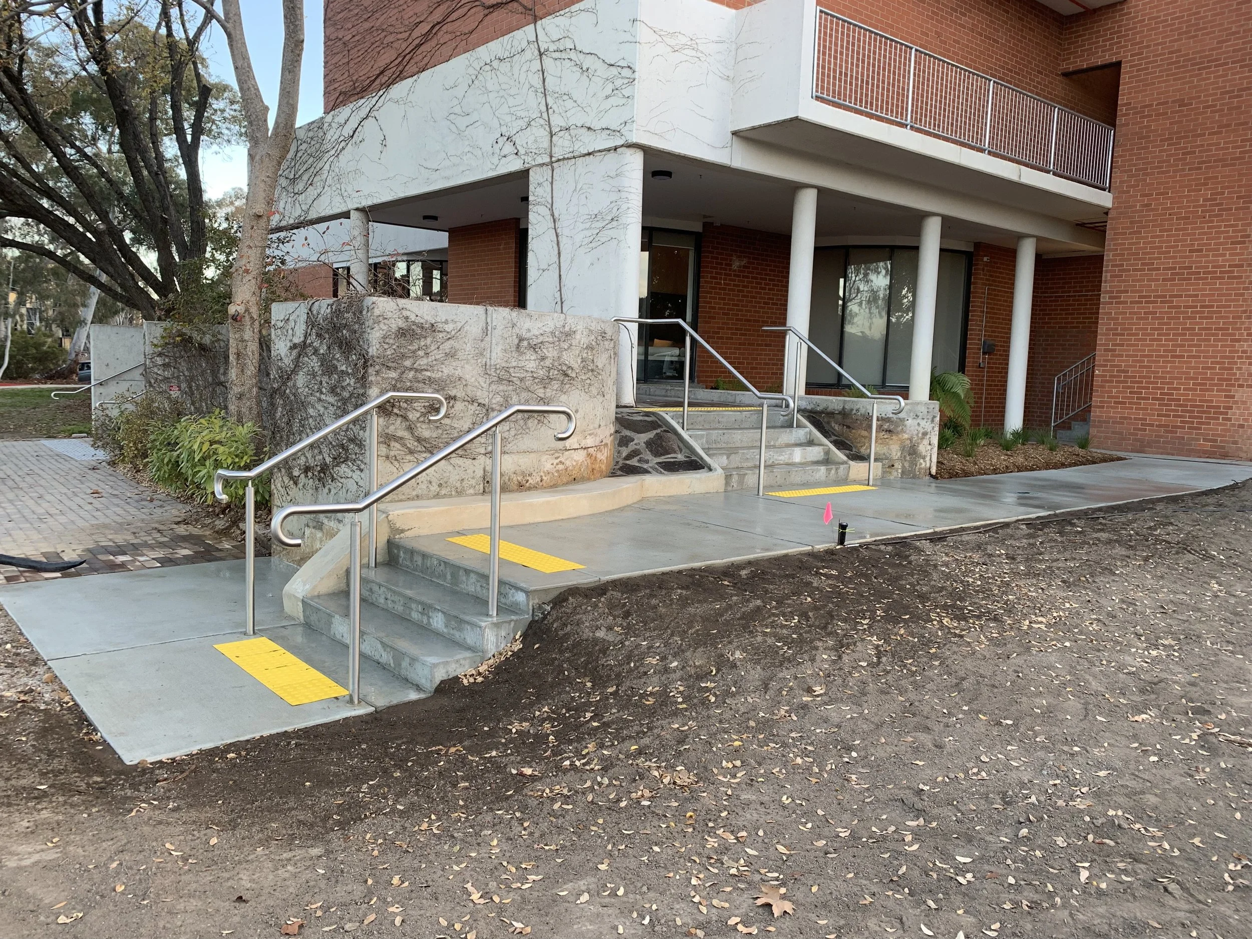 Newly constructed wheelchair accessible ramp with yellow tactile warning strips at entrance of a brick apartment building, featuring stairs and handrails, with a landscaped area and trees nearby.