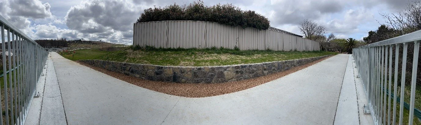 Wide-angle view of a concrete pathway with metal railings on both sides, leading to a raised grassy area with bushes and a white fence hedge, under a cloudy sky.