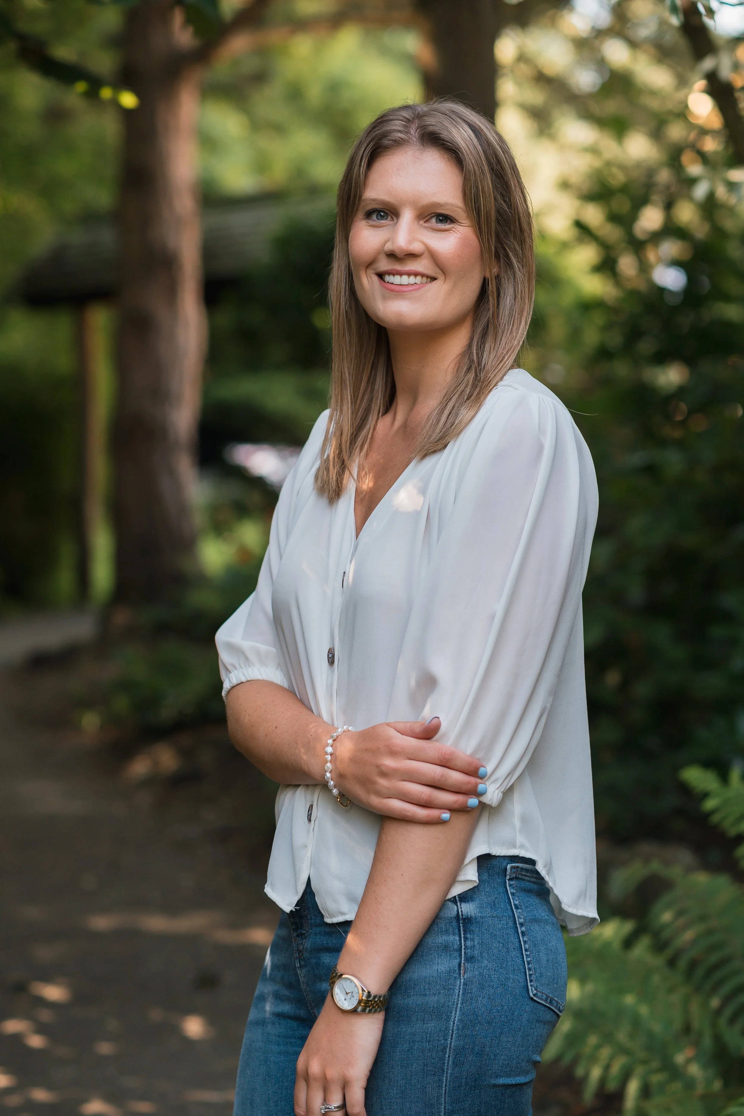 A woman in a white blouse and jeans standing outdoors in a wooded area, smiling at the camera.