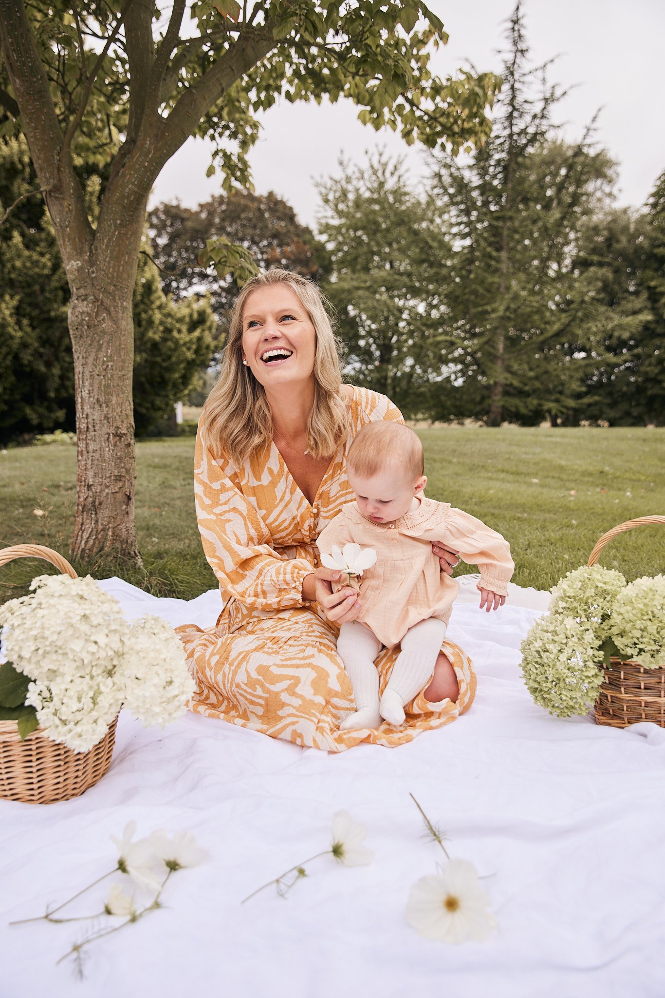 A woman and a baby sitting on a white blanket outdoors under a tree, with flowers and baskets around them; the woman is smiling and holding a flower, the baby is looking down, wearing a peach-colored dress and white tights.