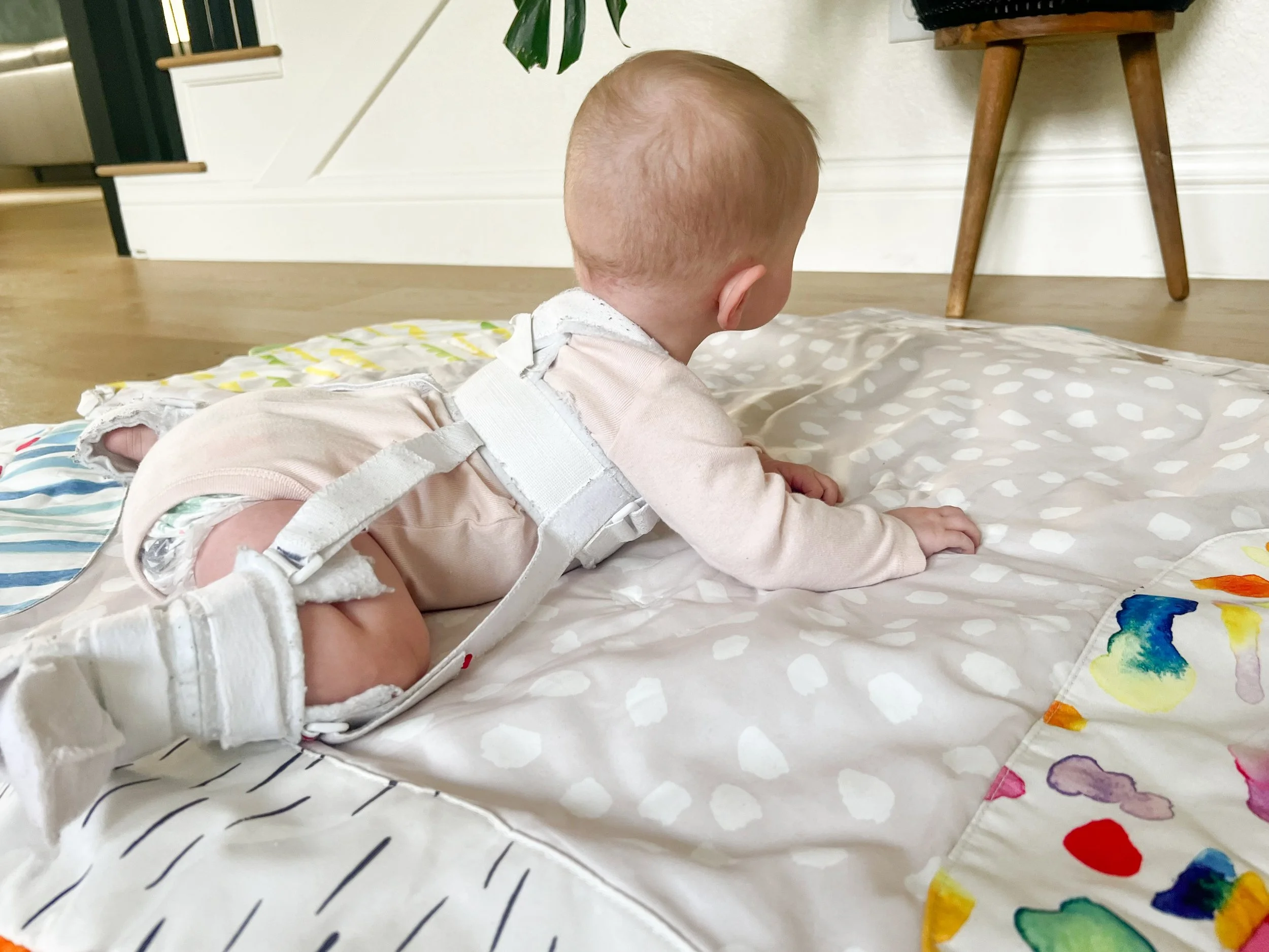 Baby lying on back on a polka dot play mat wearing a Pavlik harness, hips held in the flexed and abducted position to support healthy hip socket development
