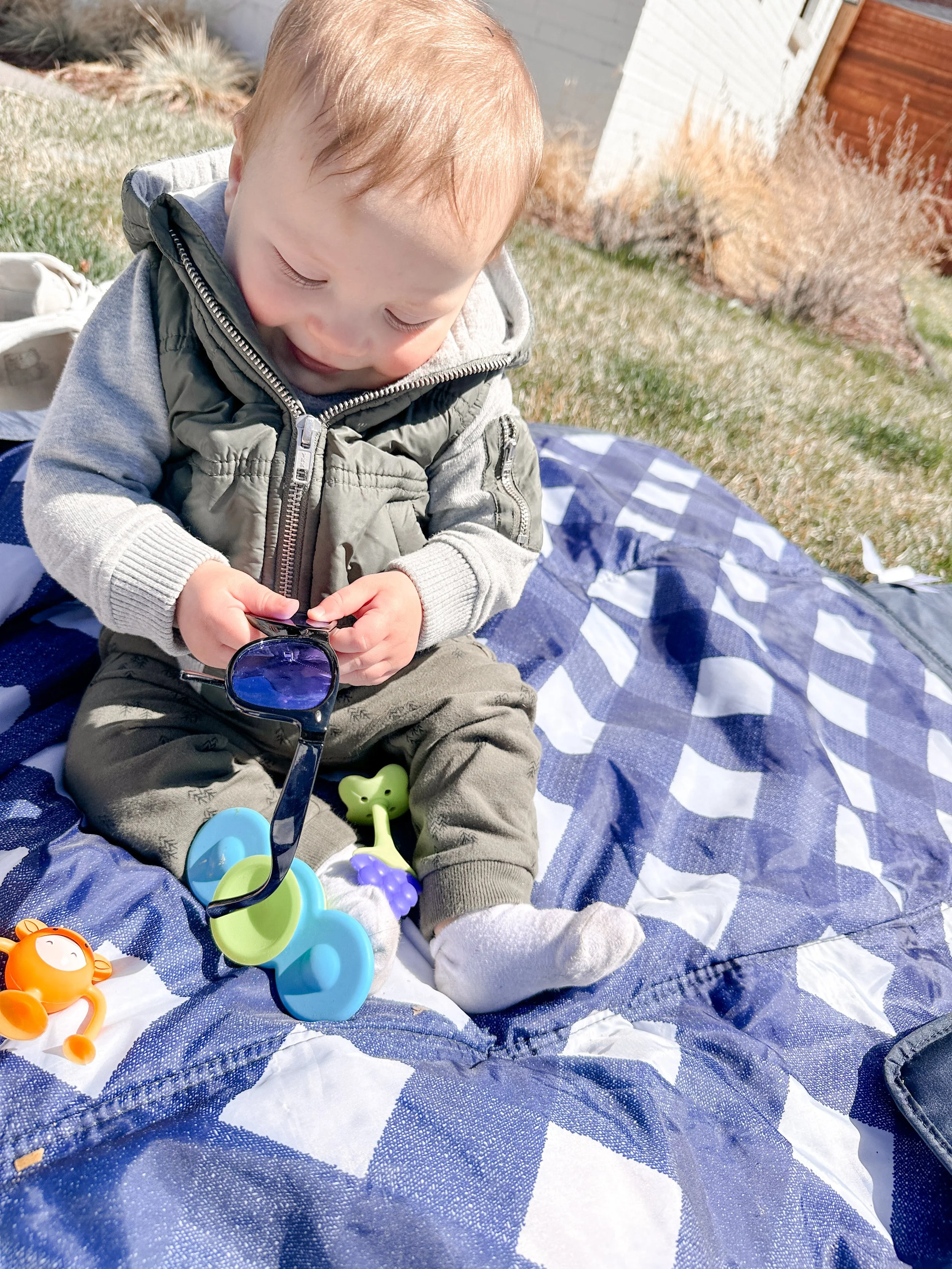 Baby sitting independently on a blanket outdoors, reaching for toys including sunglasses and sensory objects, demonstrating active floor-based sitting with reach