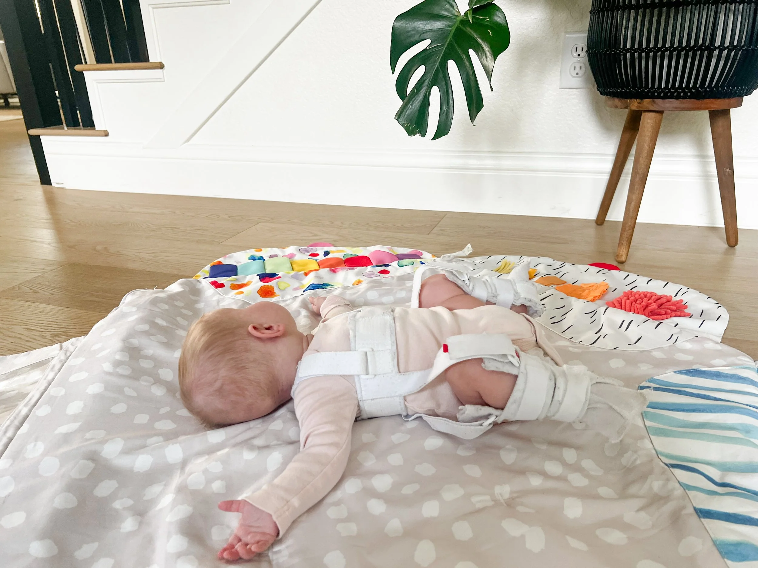 Baby lying on back on a polka dot play mat wearing a Pavlik harness, hips held in the flexed and abducted position to support healthy hip socket development