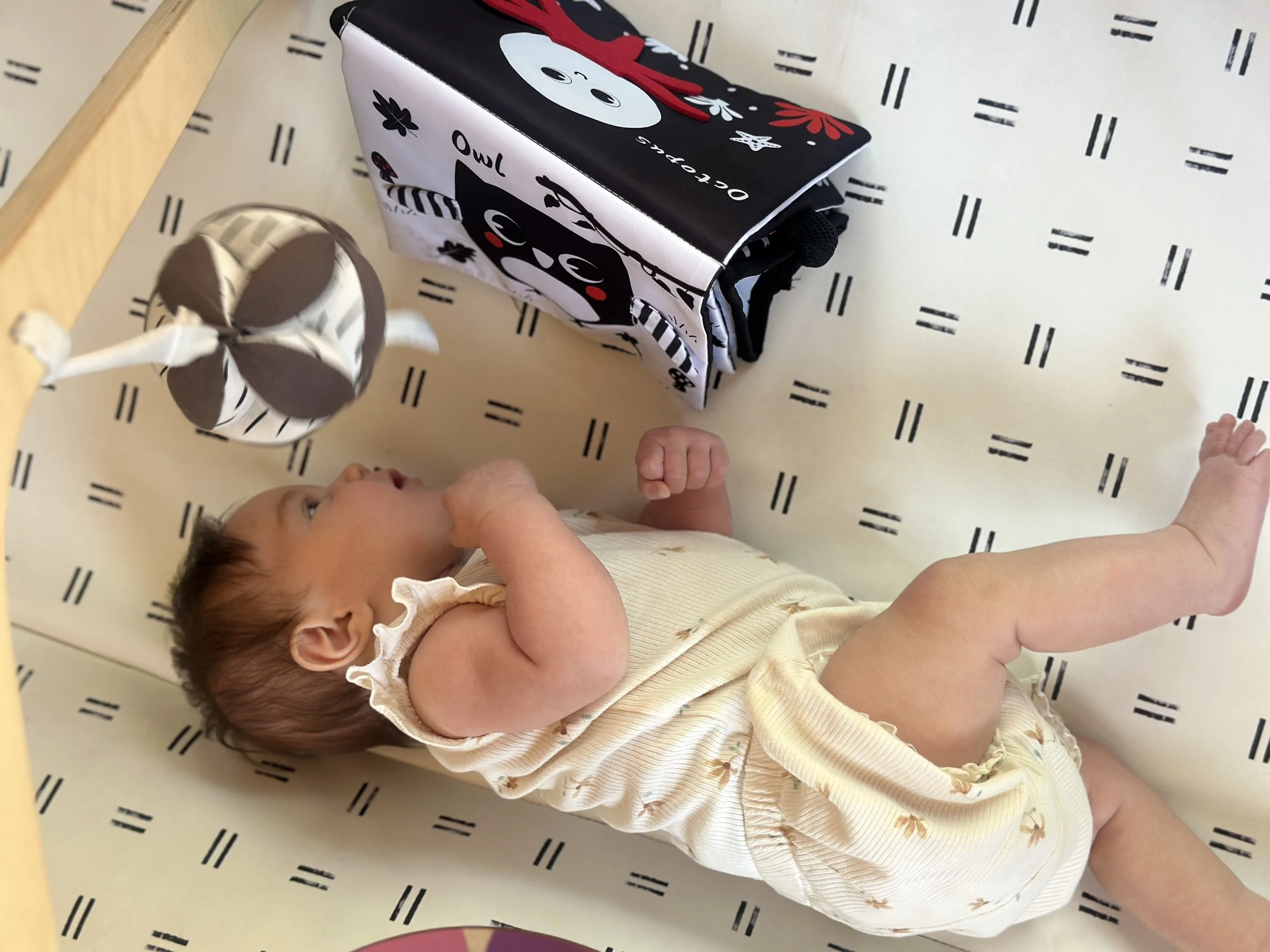 Baby on her side on a play mat reaching toward high-contrast black and white toys, practicing the weight shifting and transitional movement that builds toward rolling