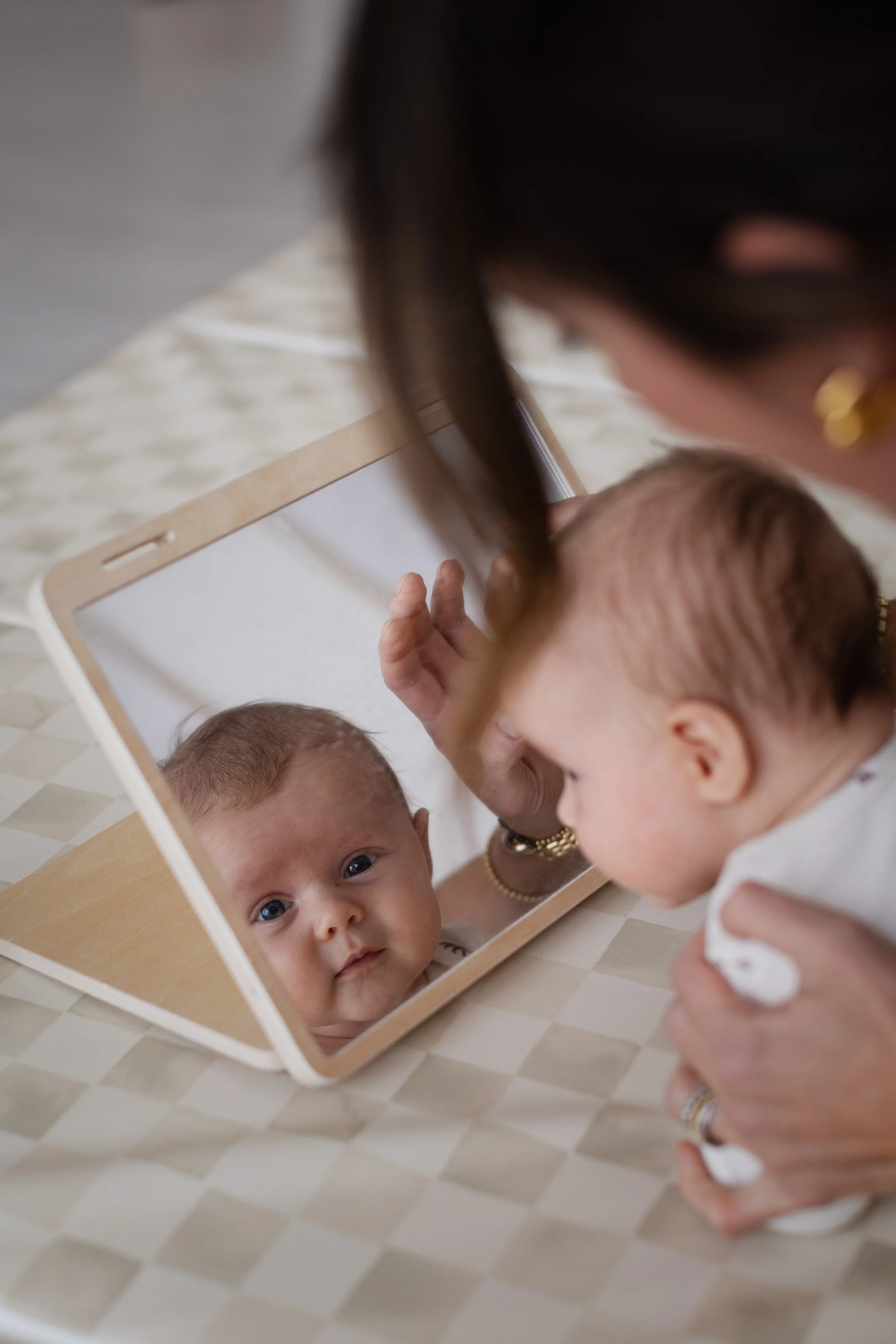 A baby looking into a mirror placed on a checkered tablecloth, with an adult holding the baby and gazing at the reflection.