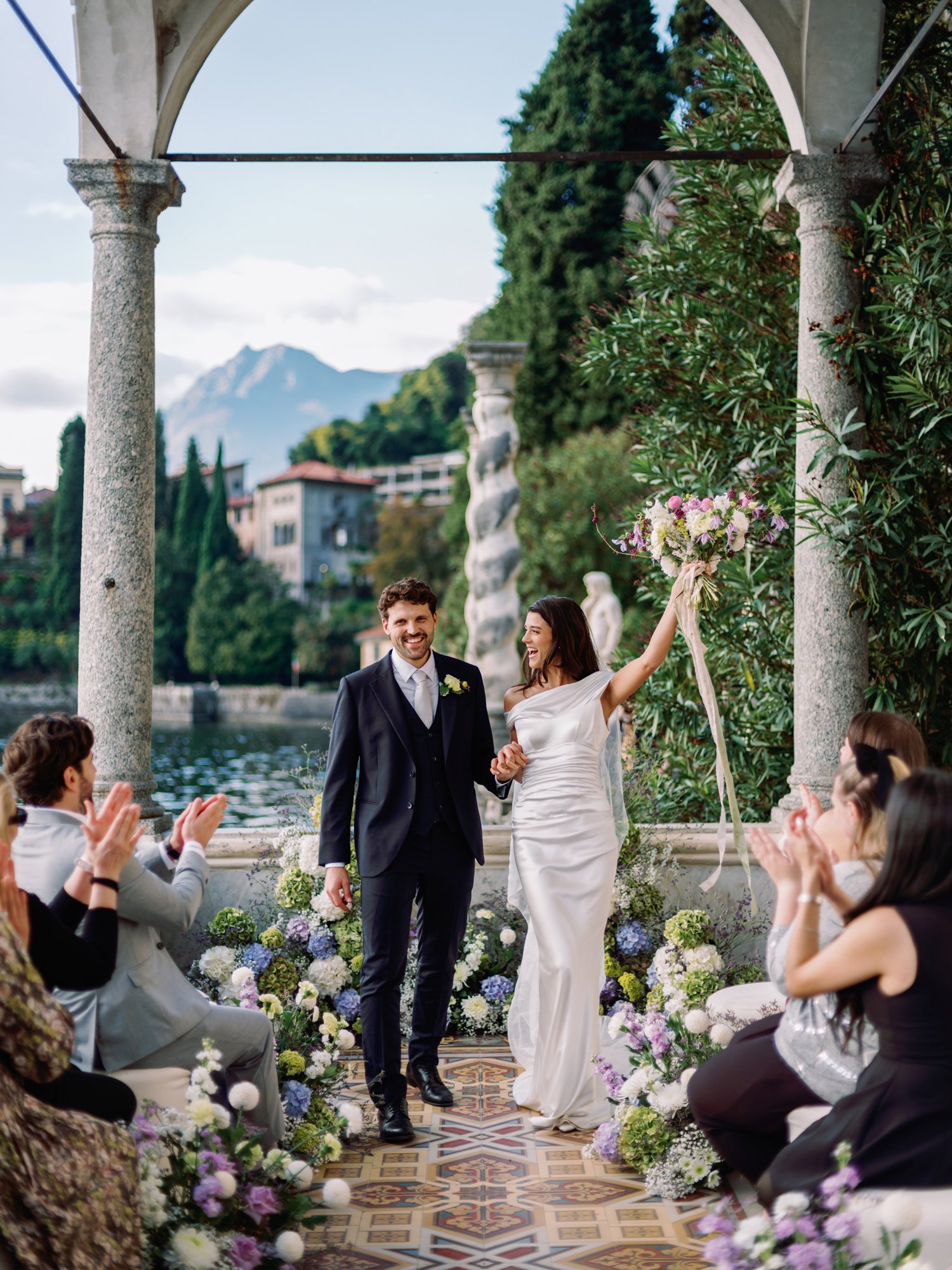 Bride and groom walking down the aisle after their ceremony at Villa Monastero on Lake Como, Italy, surrounded by colorful floral arrangements and applauding guests, with the lake and mountains in the background.