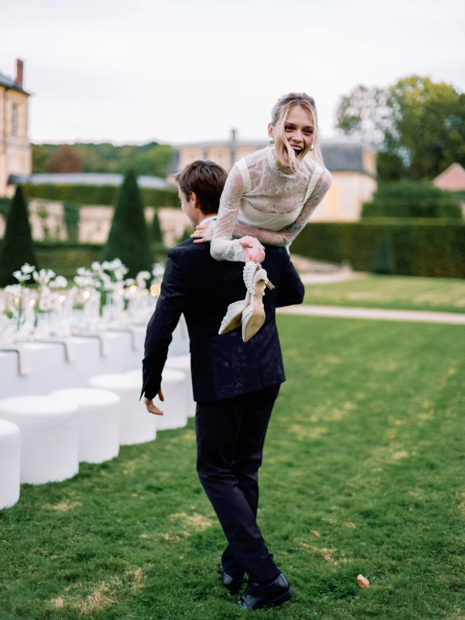 Bride laughing as the groom carries her through the gardens during a wedding at Château de Villette in France, holding her shoes in one hand, with elegant ceremony seating, glowing candles, and manicured château grounds in the background.