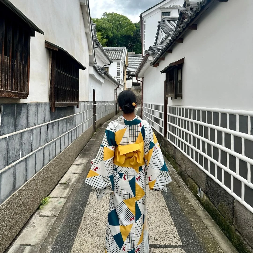 Founder of Worthy Wanderlust in a kimono, standing in a narrow alley in the Bikan Historical Quarter  (white houses with namako tiles).