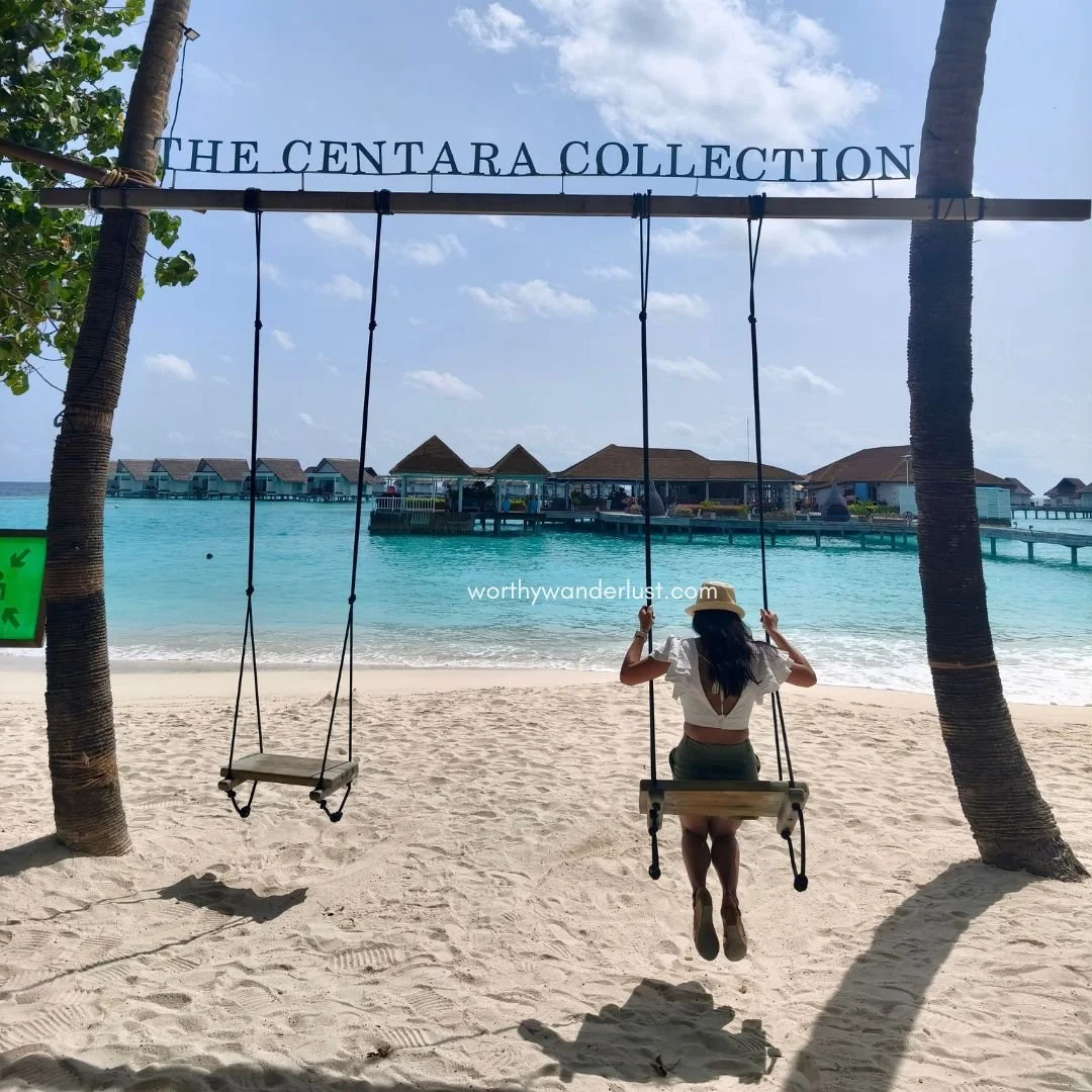 Lady sitting on a swing, with 'The Centara Collection' above it, facing overwater villas