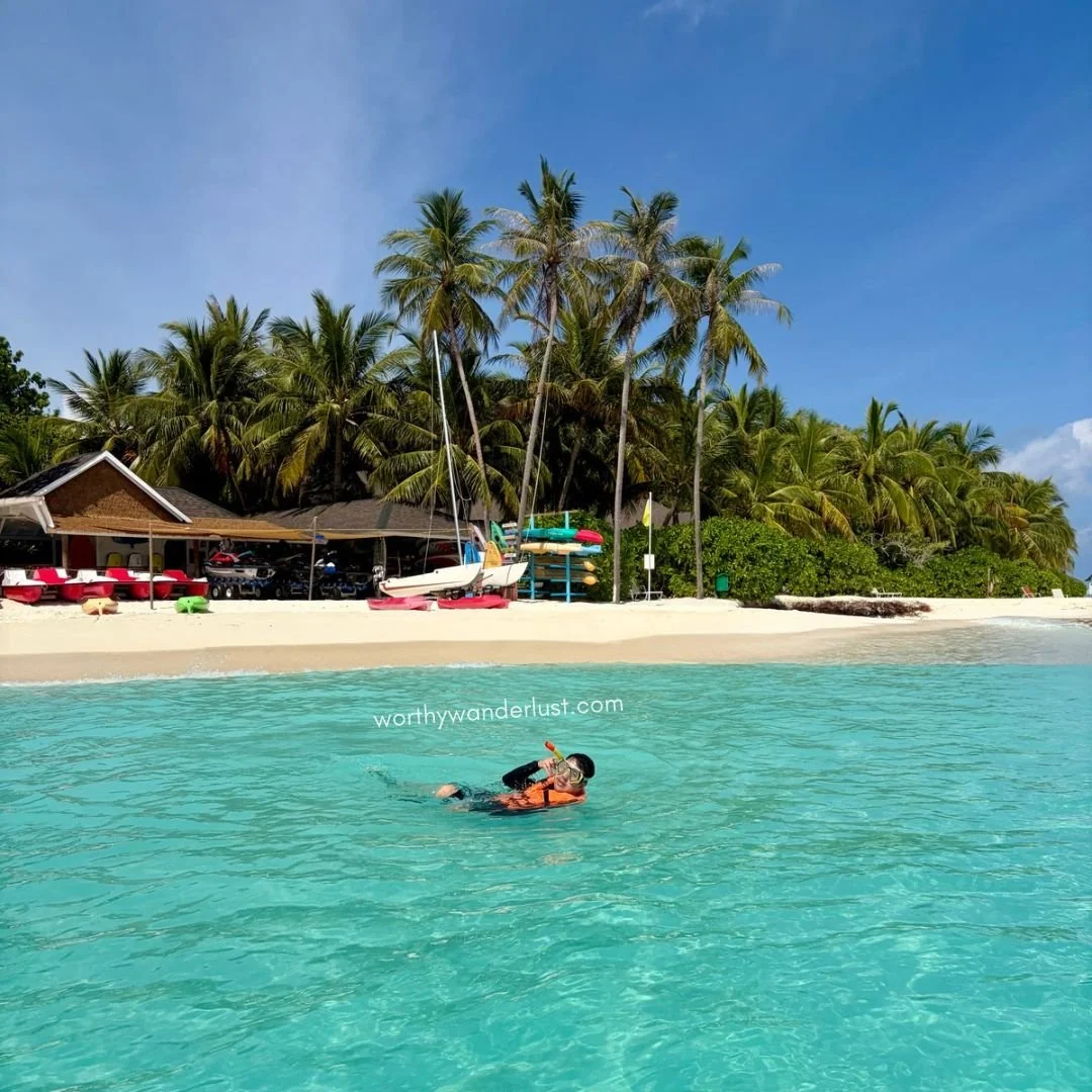 Man in clear turqoise water, in front of a beach adn water sports centre, with many palm trees.
