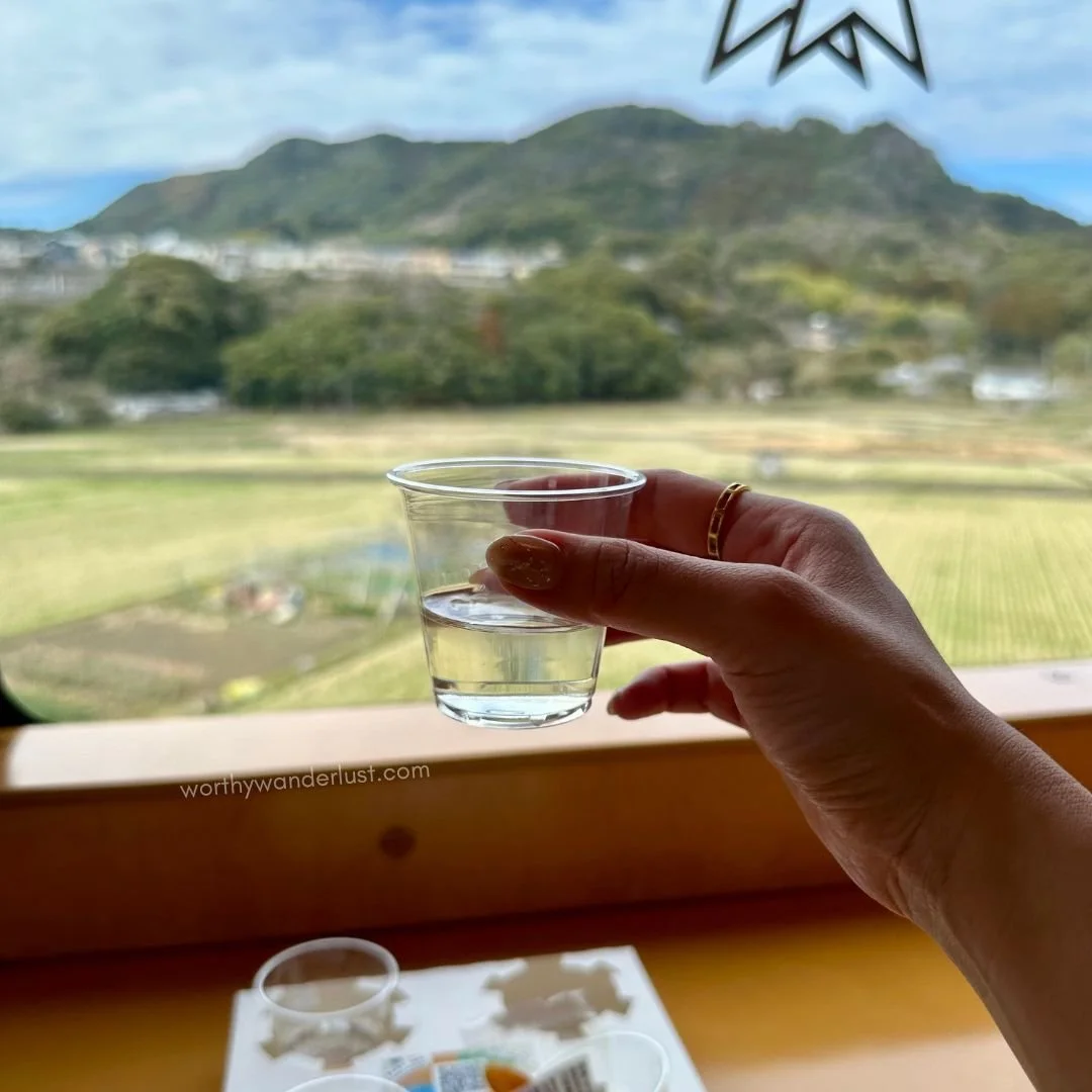 Hand holding a small cup of sake against a window showing paddy fields and greenery.