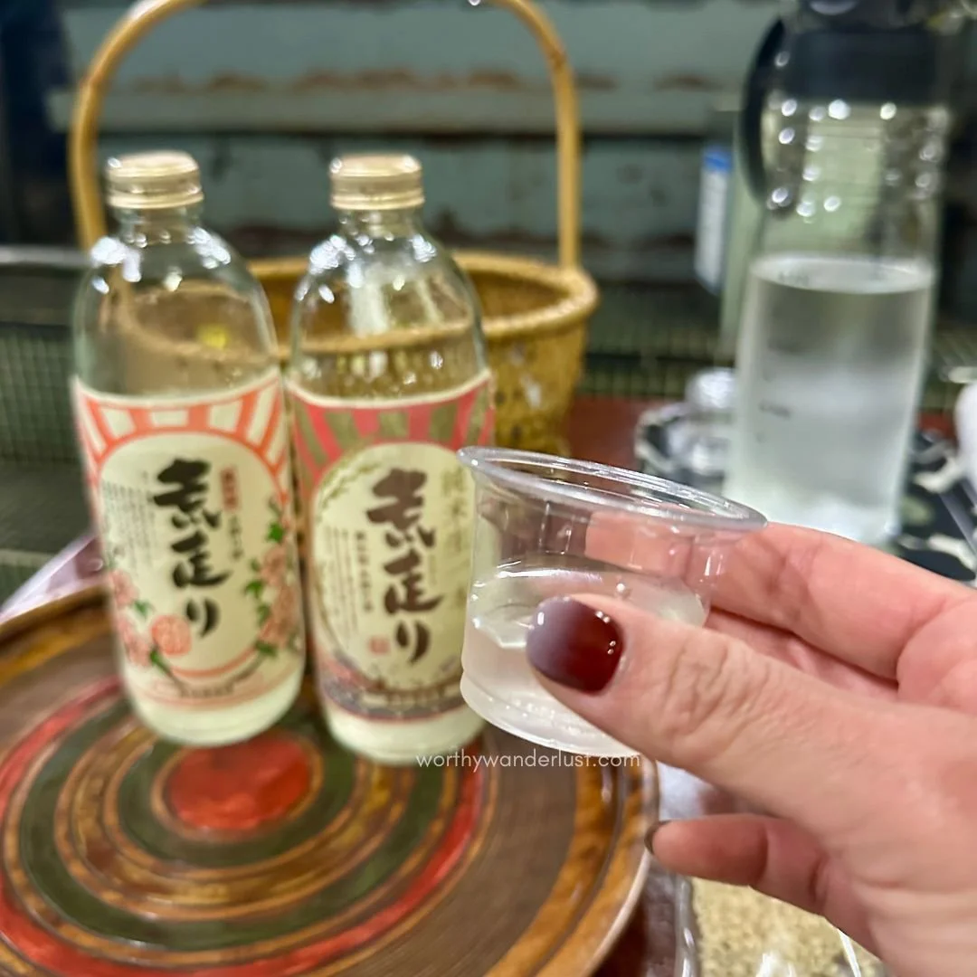 Hand holding up a sample of sake with sake bottles in the background