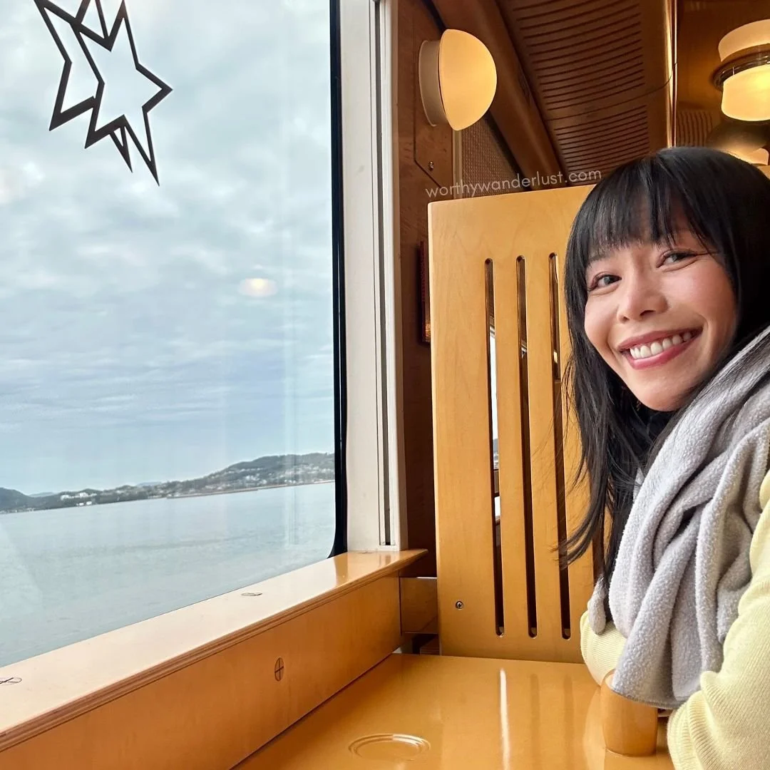 Worthywanderlust.com founder smiling at a seat facing a large window with sea-views