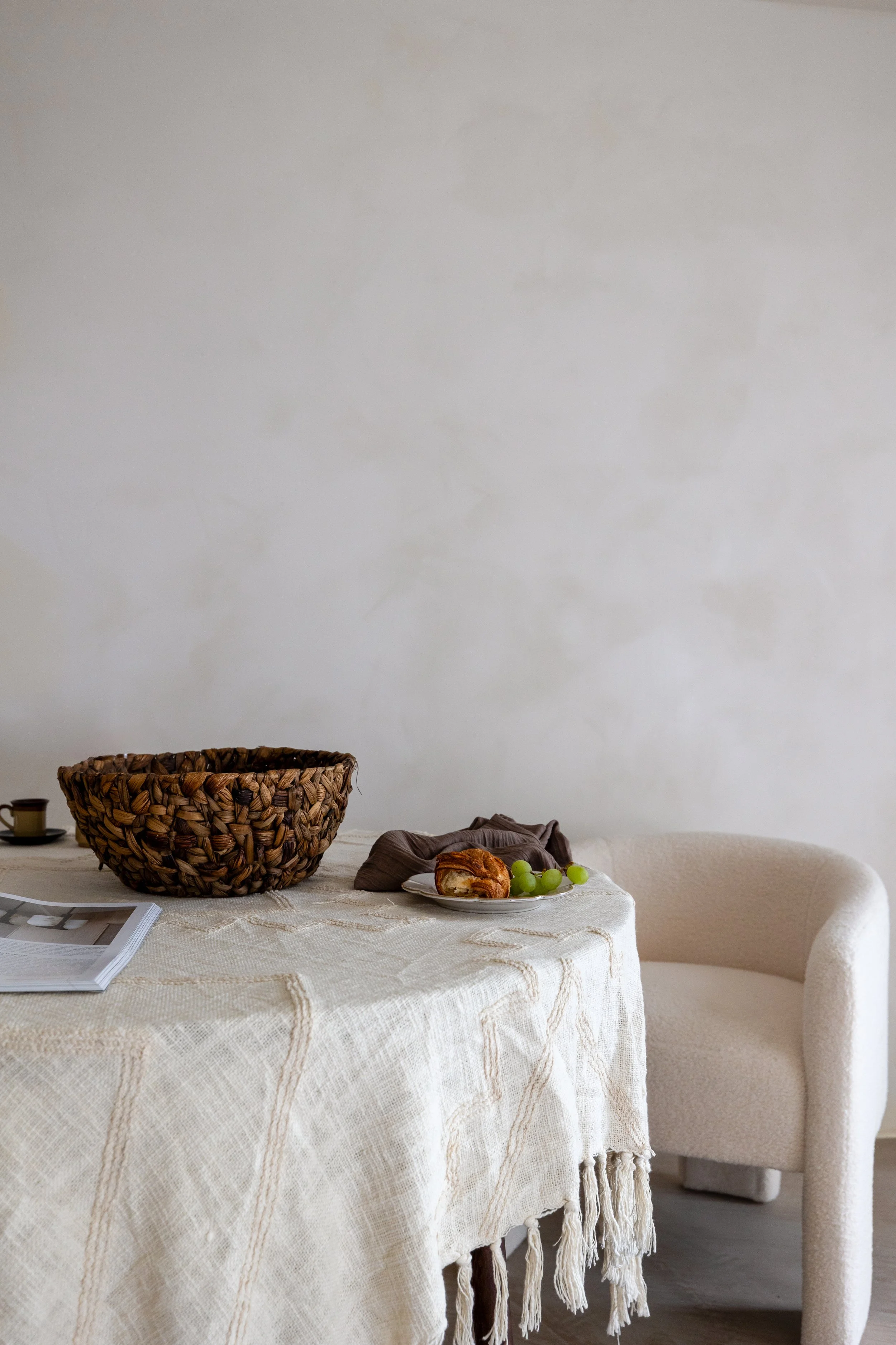 Cozy dining setting with a round table, a white fringed tablecloth, a wicker basket, a plate with a croissant and green grapes, a cup, and an open magazine.