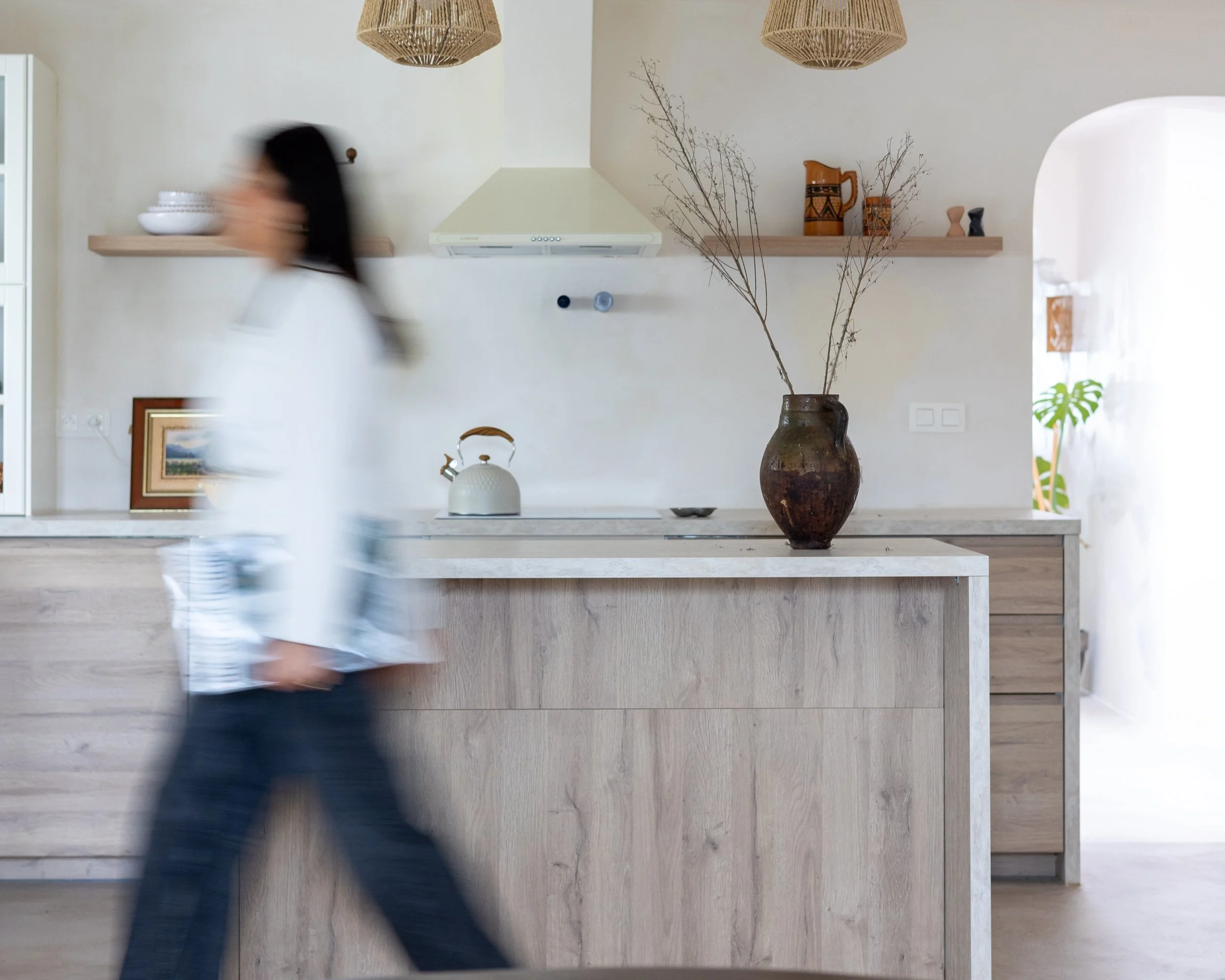 Modern kitchen interior with wooden cabinets, a countertop, a rustic vase with branches, a blurry person walking by, and decorative items on shelves. A white range hood and kettle are visible.
