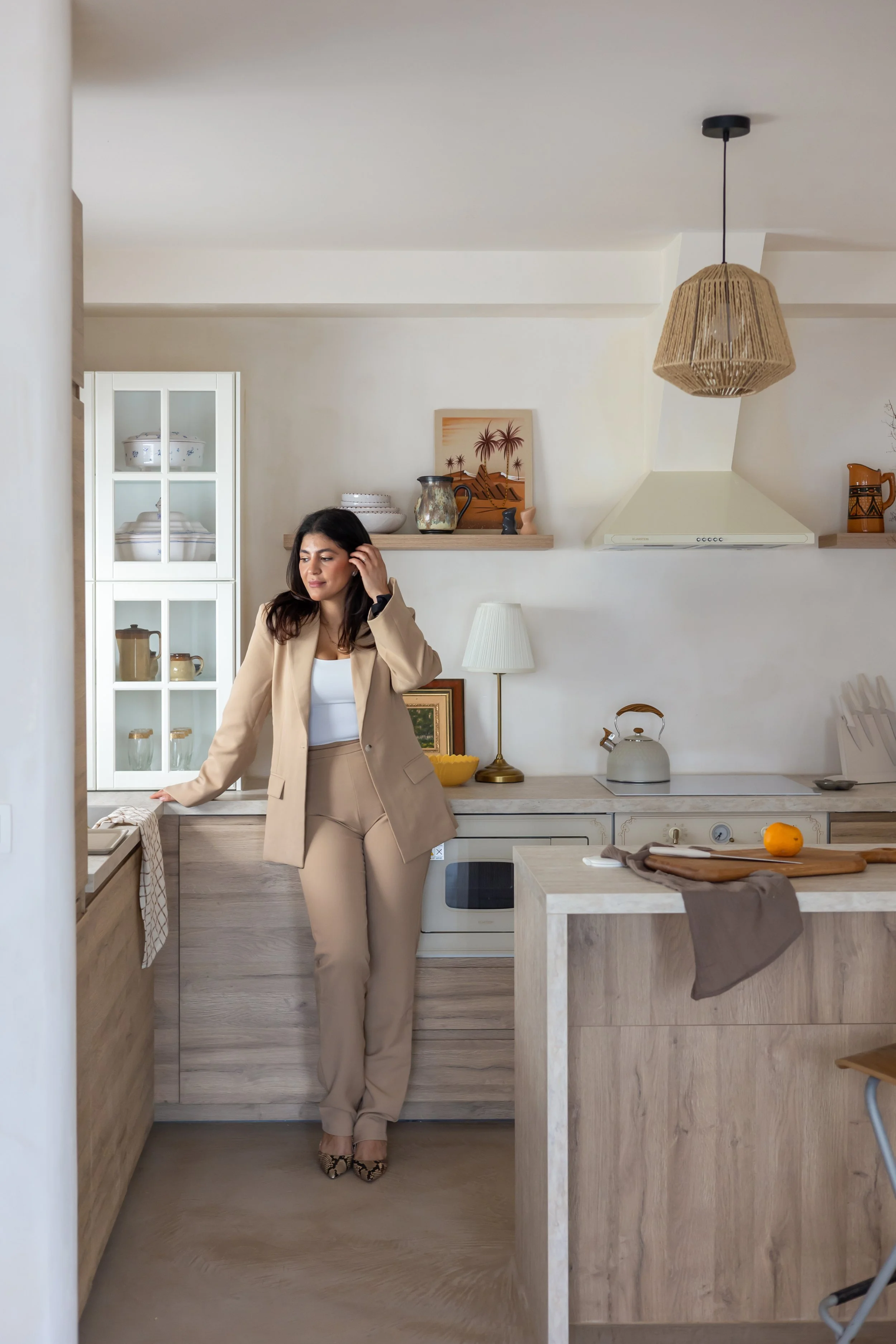 Woman in beige suit standing in a modern kitchen with wooden cabinets, a white countertop, and decorative items on shelves.