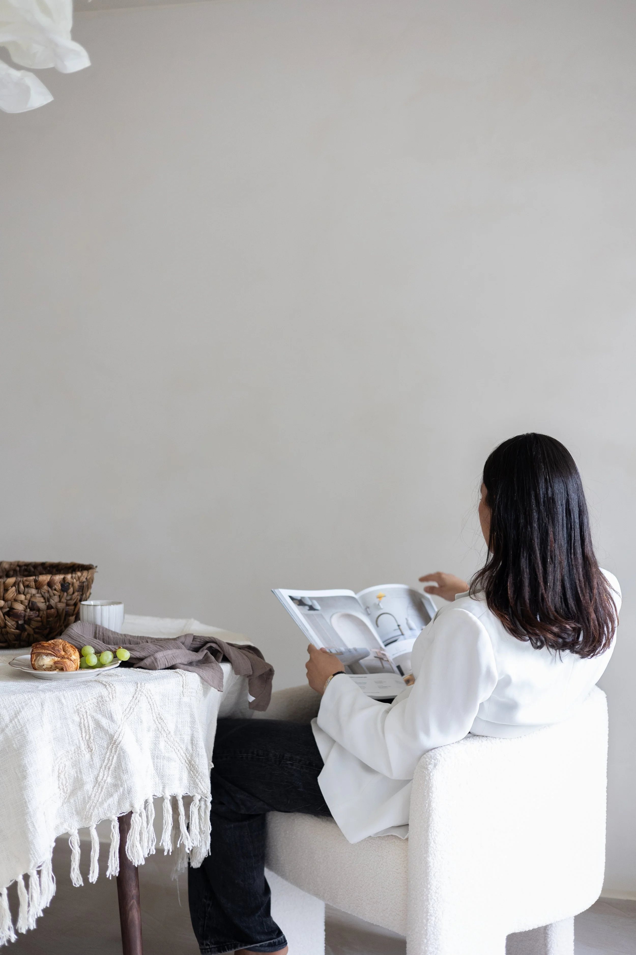 Person in a white chair reading a magazine at a table with a white tablecloth, a plate with a croissant and grapes, and a wicker basket.