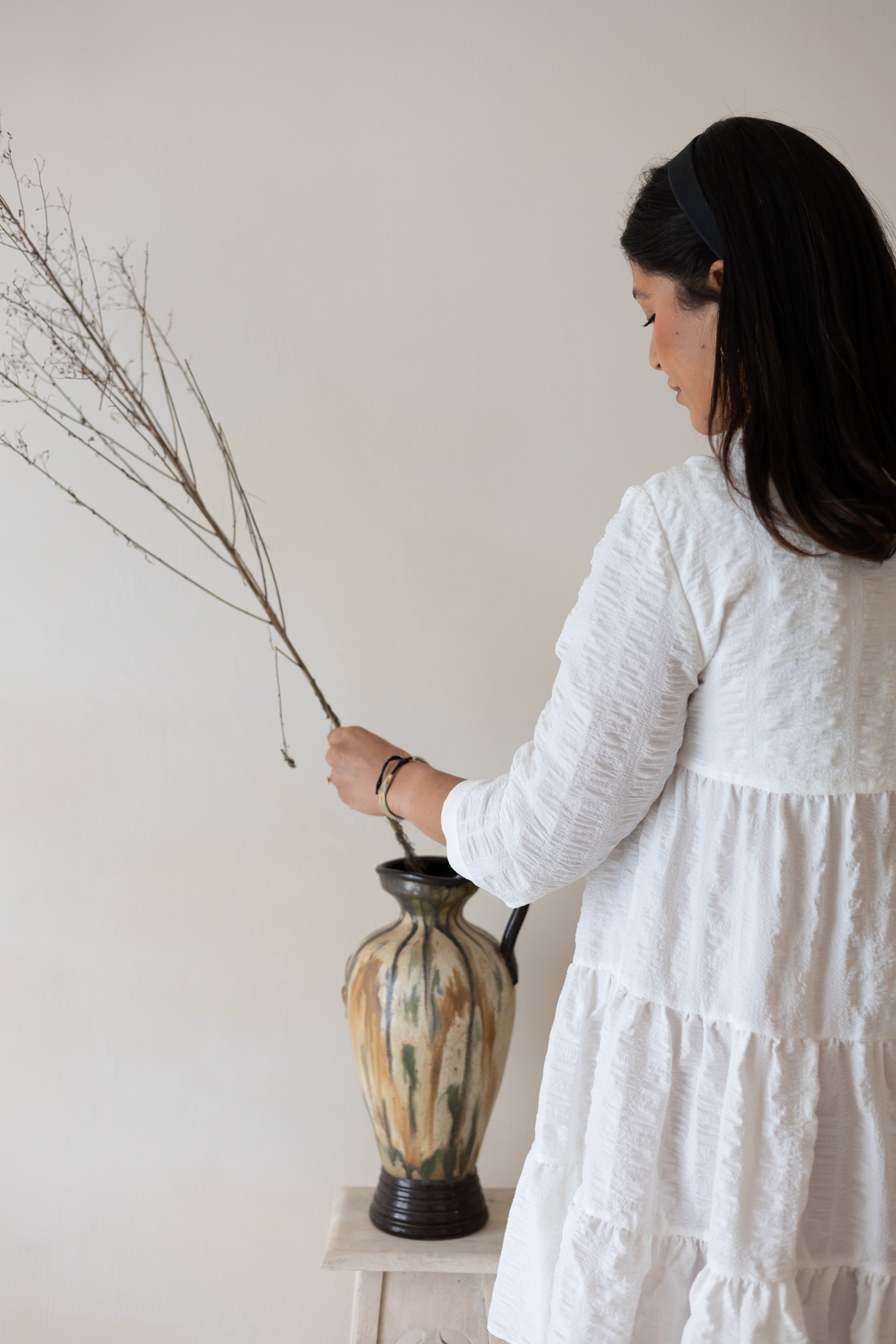 Woman in white dress arranging dried branches in a decorative vase on a wooden table.