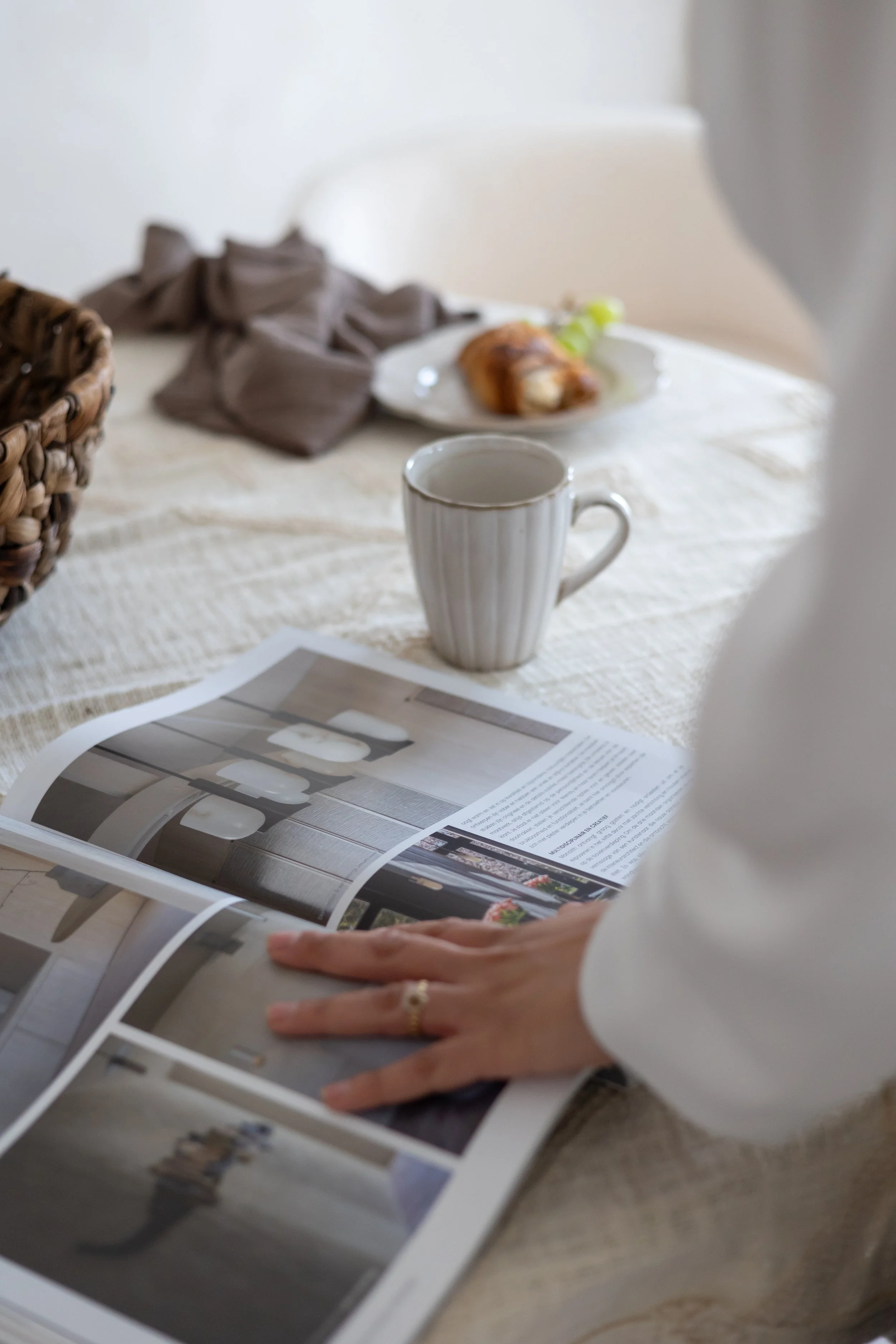 Person reading a magazine with a cup of tea or coffee on a table, alongside a plate with a pastry.