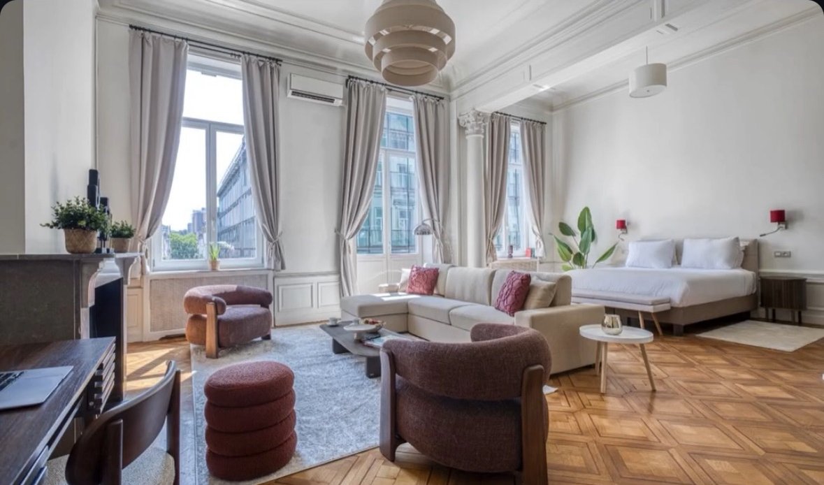 Living room with large windows, beige curtains, a white sofa with red pillows, a bed in the background, a round white coffee table, and pink and purple chairs, with hardwood floors and modern decor.