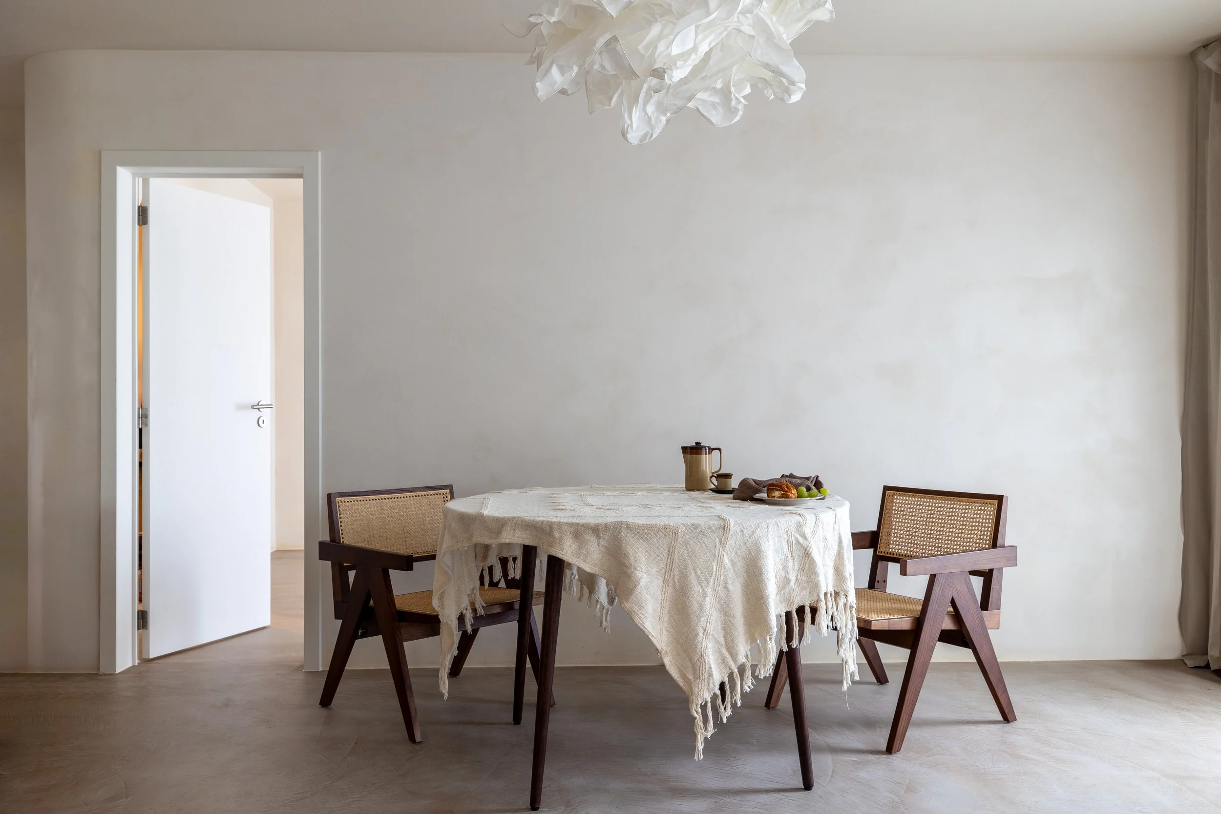 Minimalist dining room with a round table covered by a white fringed tablecloth, surrounded by three wooden chairs with woven seats. A light fixture resembling a cloud hangs above. A doorway is open to the left.