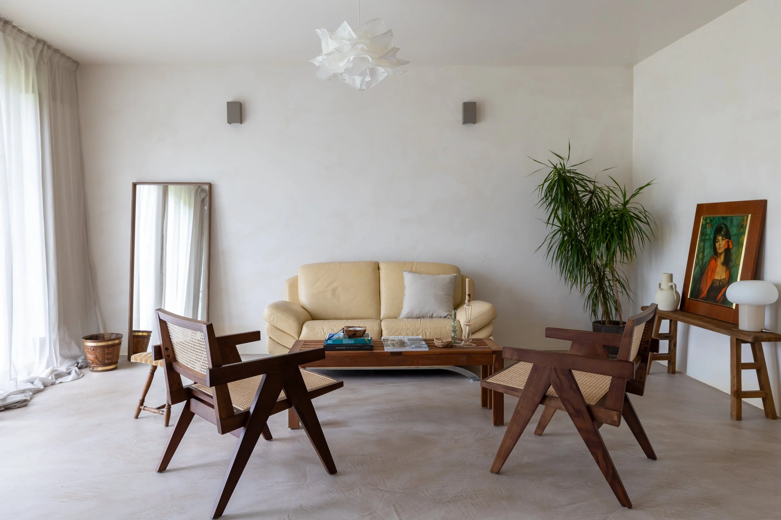 Minimalist living room with a beige sofa, two wooden chairs, a wooden coffee table, a large potted plant, a framed painting, a floor mirror, and sheer curtains.