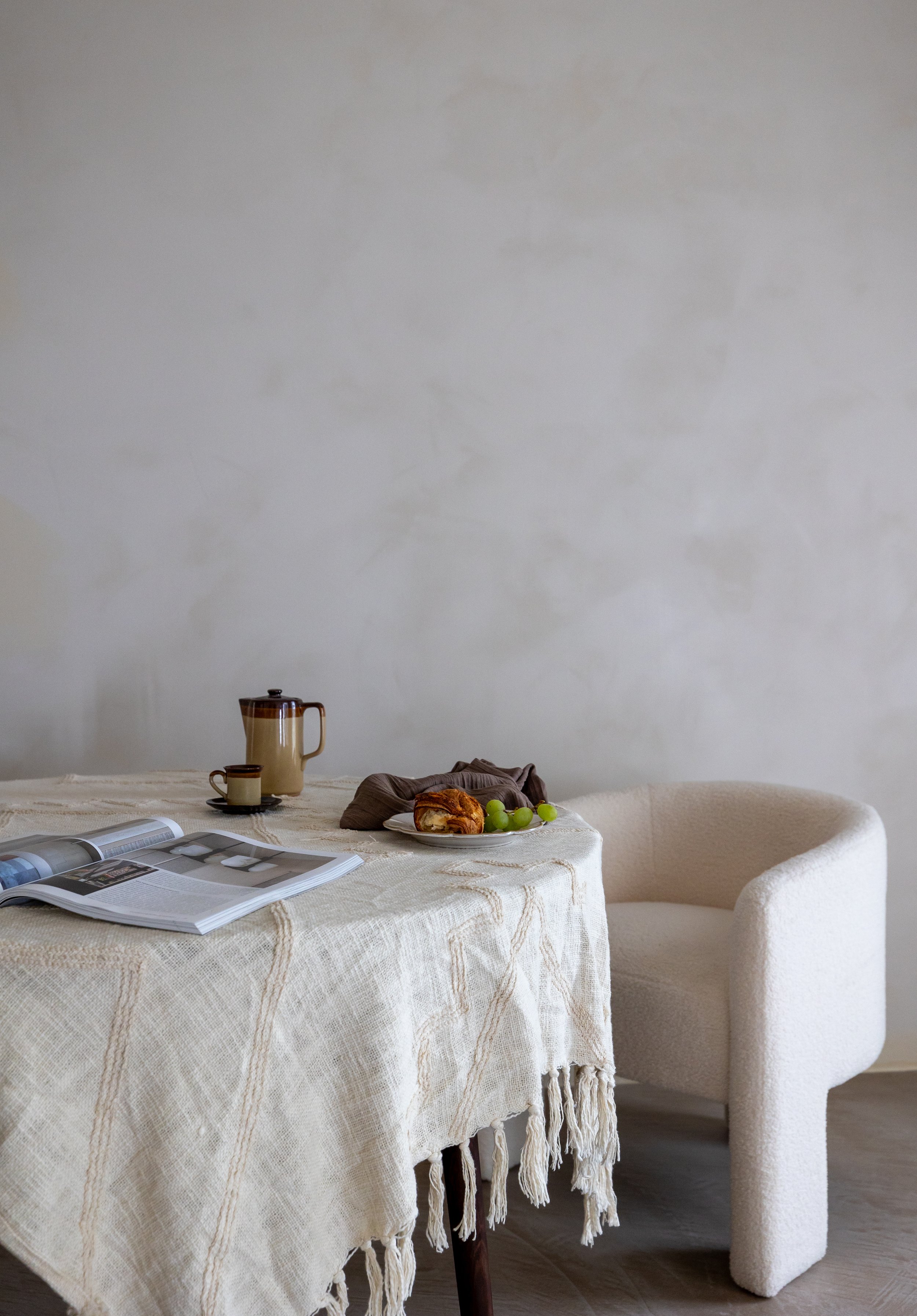 Minimalist dining scene with a round table covered by a beige textured tablecloth, a chair, an open book, a cup, a coffee pot, and a plate with a pastry and green grapes.